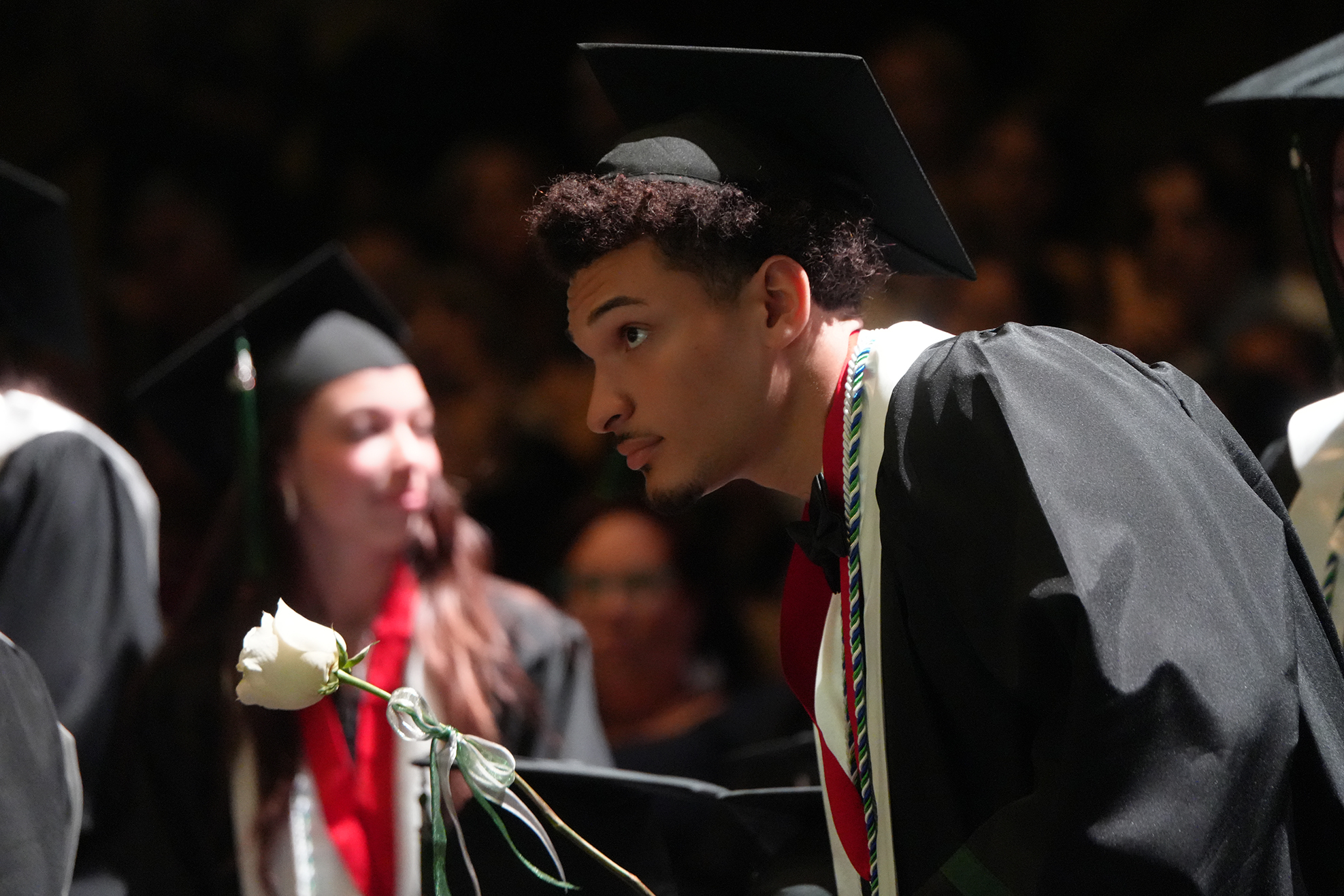 A young man in a graduation gown, holding a white flower, stands at a graduation ceremony.