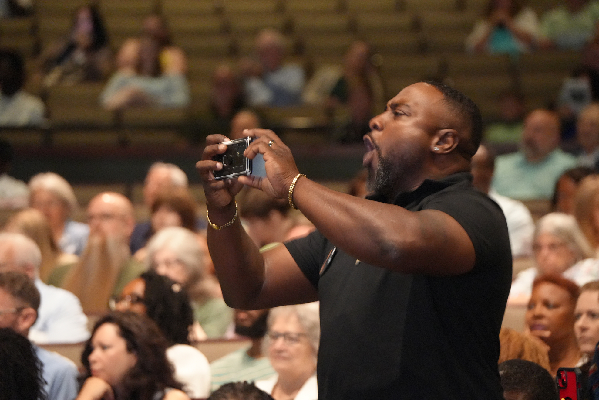A man captures a moment on his camera, standing before a crowd in a room, some seated on bleachers.