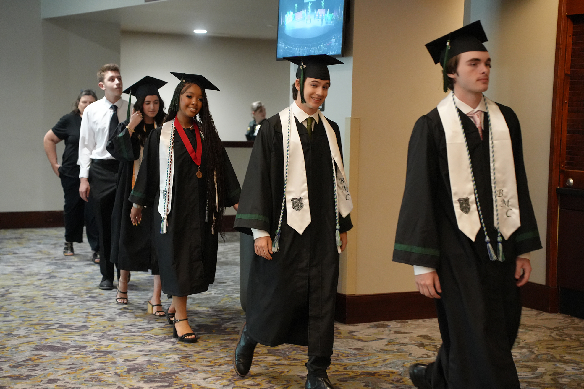 A group of people wearing graduation gowns and hats walking down a hall. A TV is on a wall.