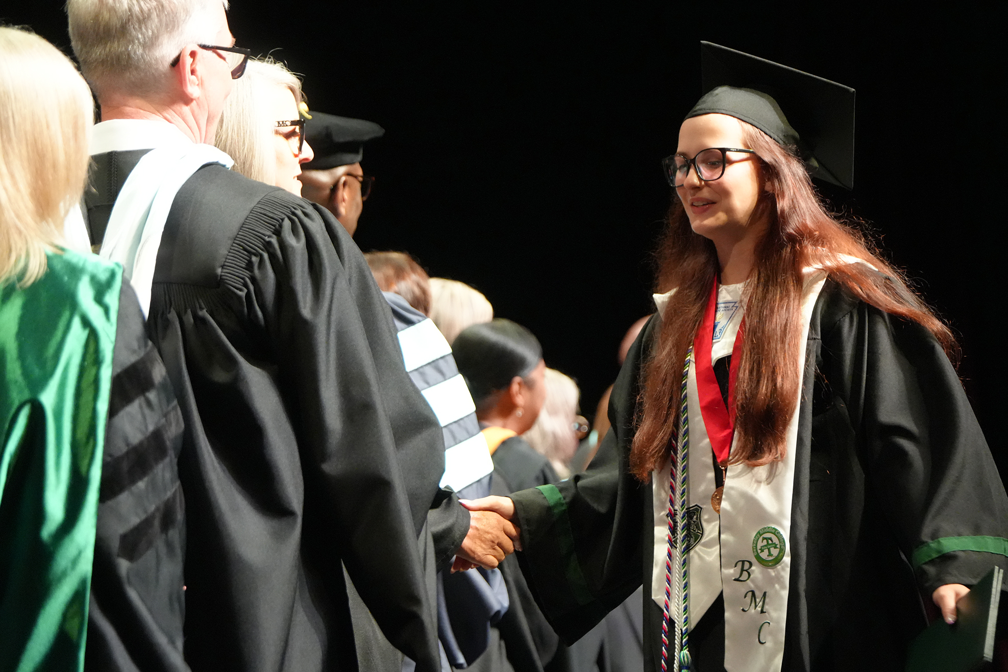 A woman in a graduation gown walks forward, shaking hands with a person in a black academic robe.