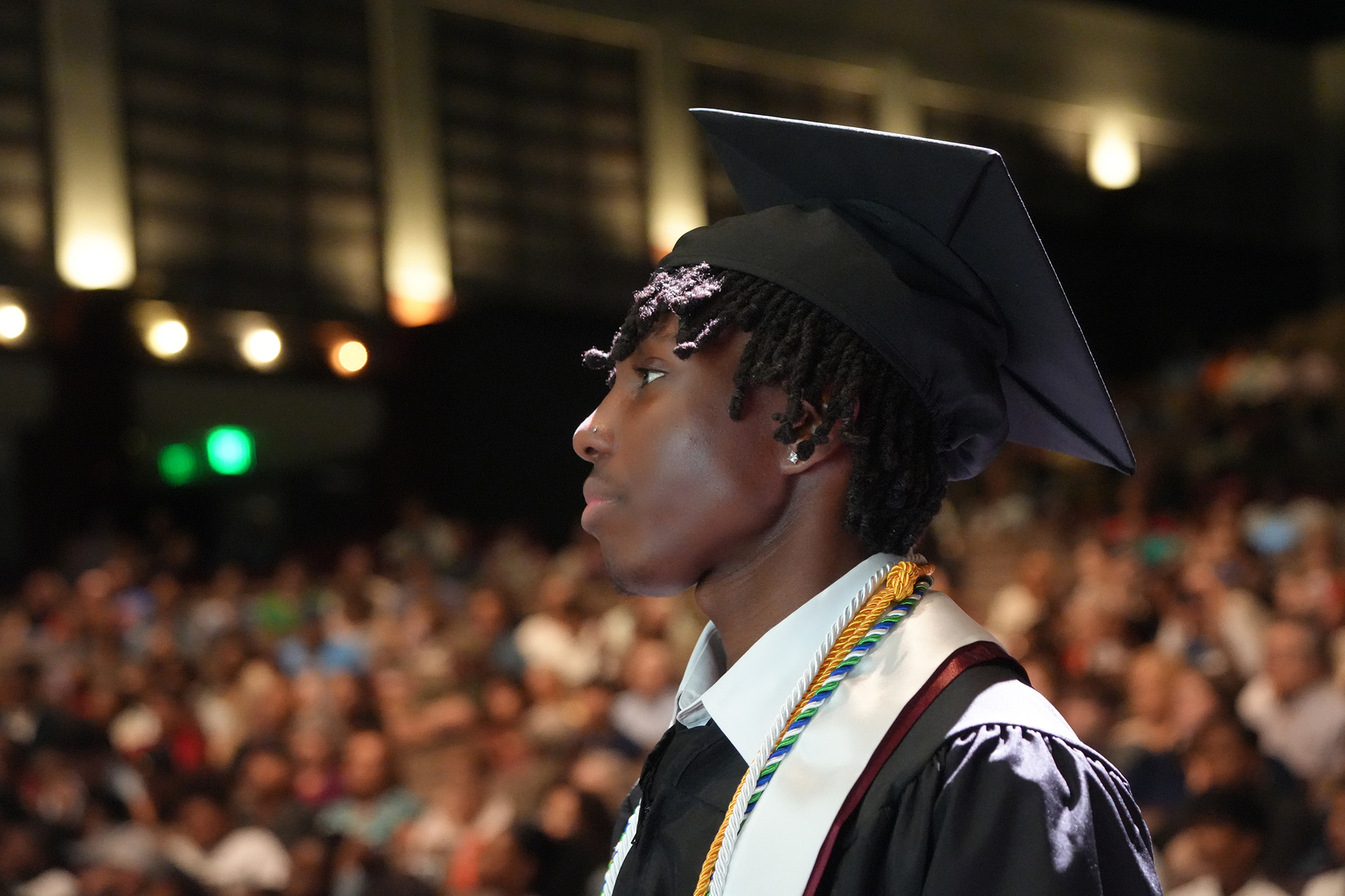 A graduate stands at an event, wearing a cap, gown, and stole, facing left, with a blurred audience in the background.