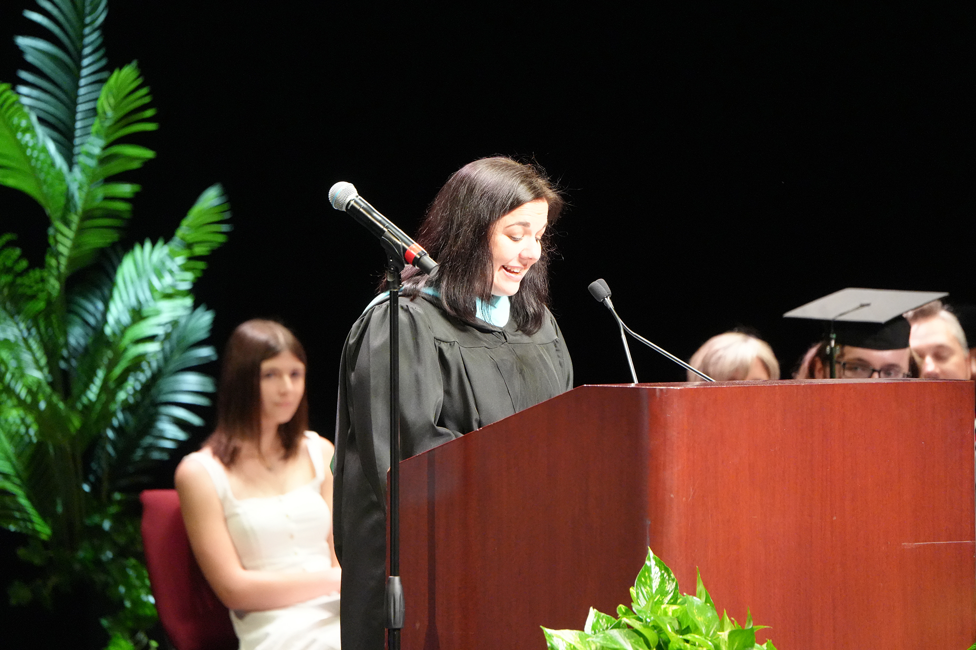 A woman stands at a podium with a microphone, wearing a graduation gown. Another woman sits behind her. A plant is visible in the background.