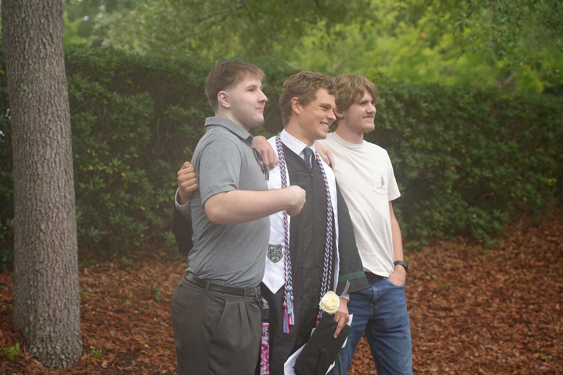 Three individuals pose together in a leafy, wooded area. Two have arms on each other's shoulders, and one wears a graduation gown.