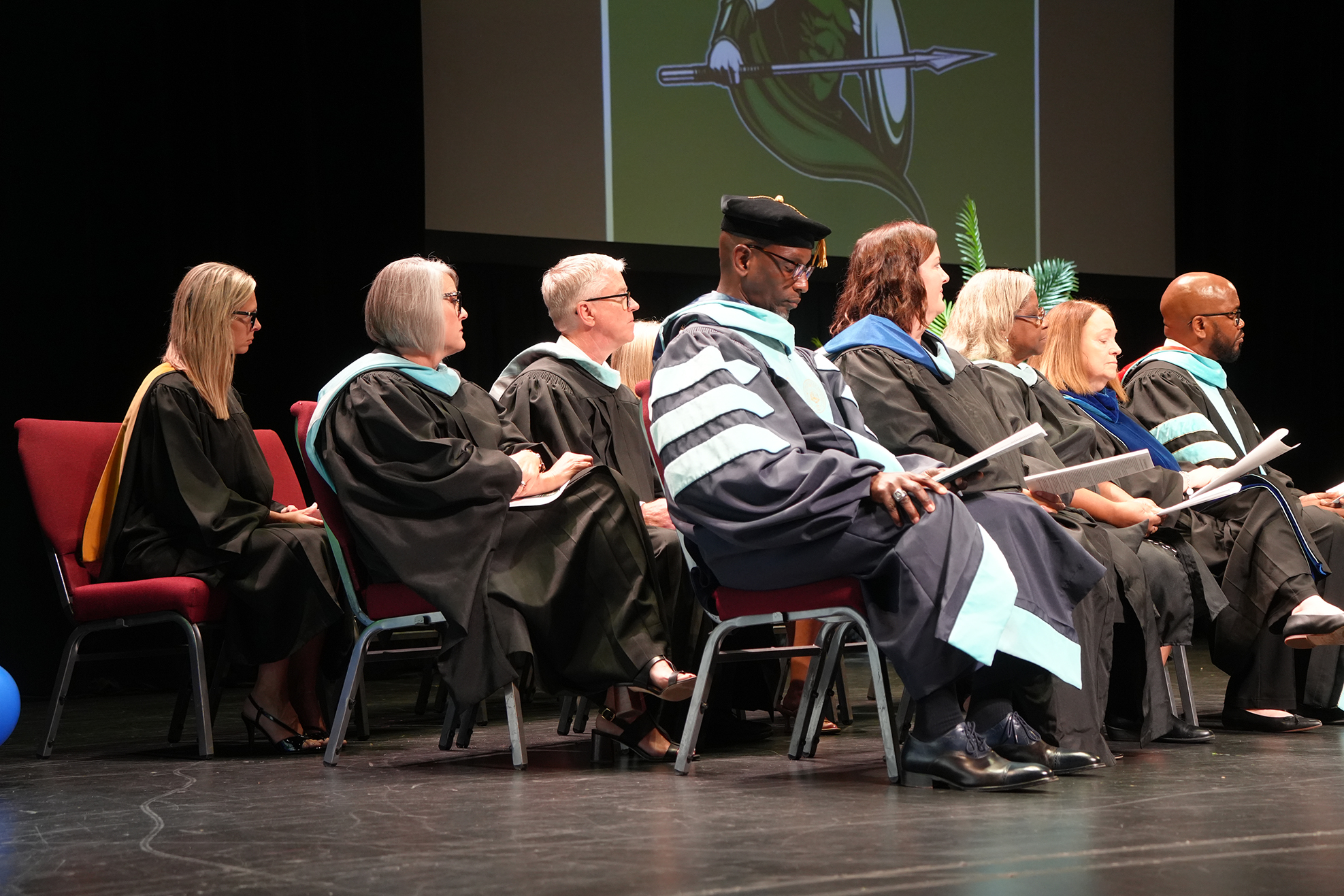 Graduates in academic gowns and caps sitting on chairs on a stage, a screen behind them.