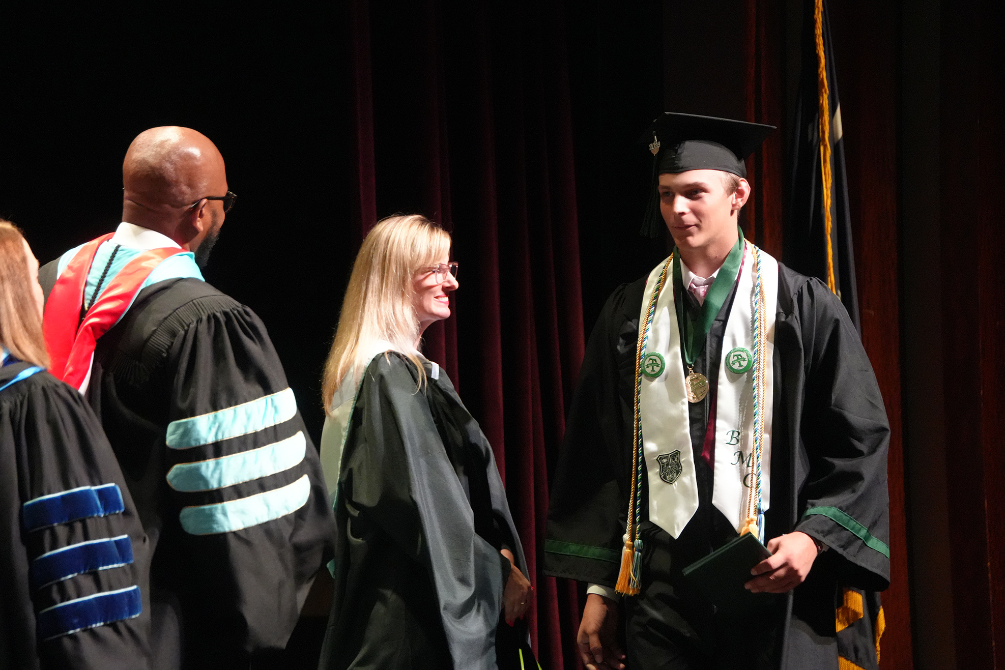 A man and woman in graduation gowns and hats stand on a stage, likely at a ceremony.