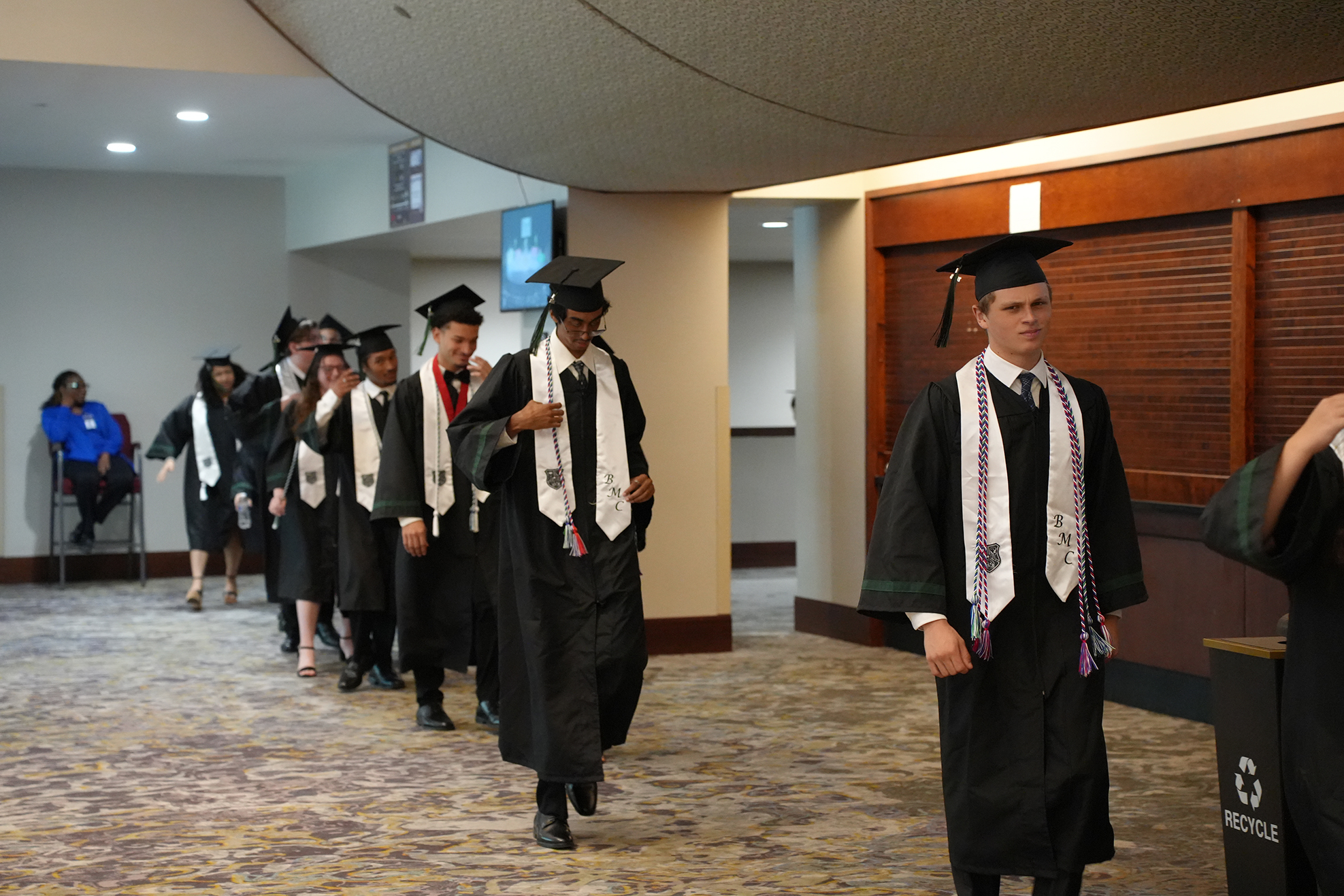 Graduates in gowns and caps walking in a corridor, a person at the end holding a bin.