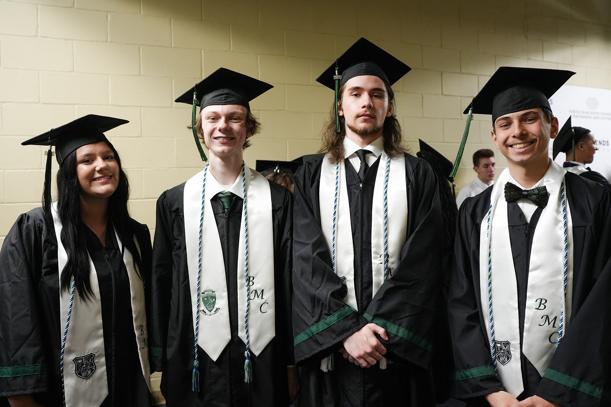 Four people in black graduation robes, hats, and sashes, posing in a hallway with a yellow wall.