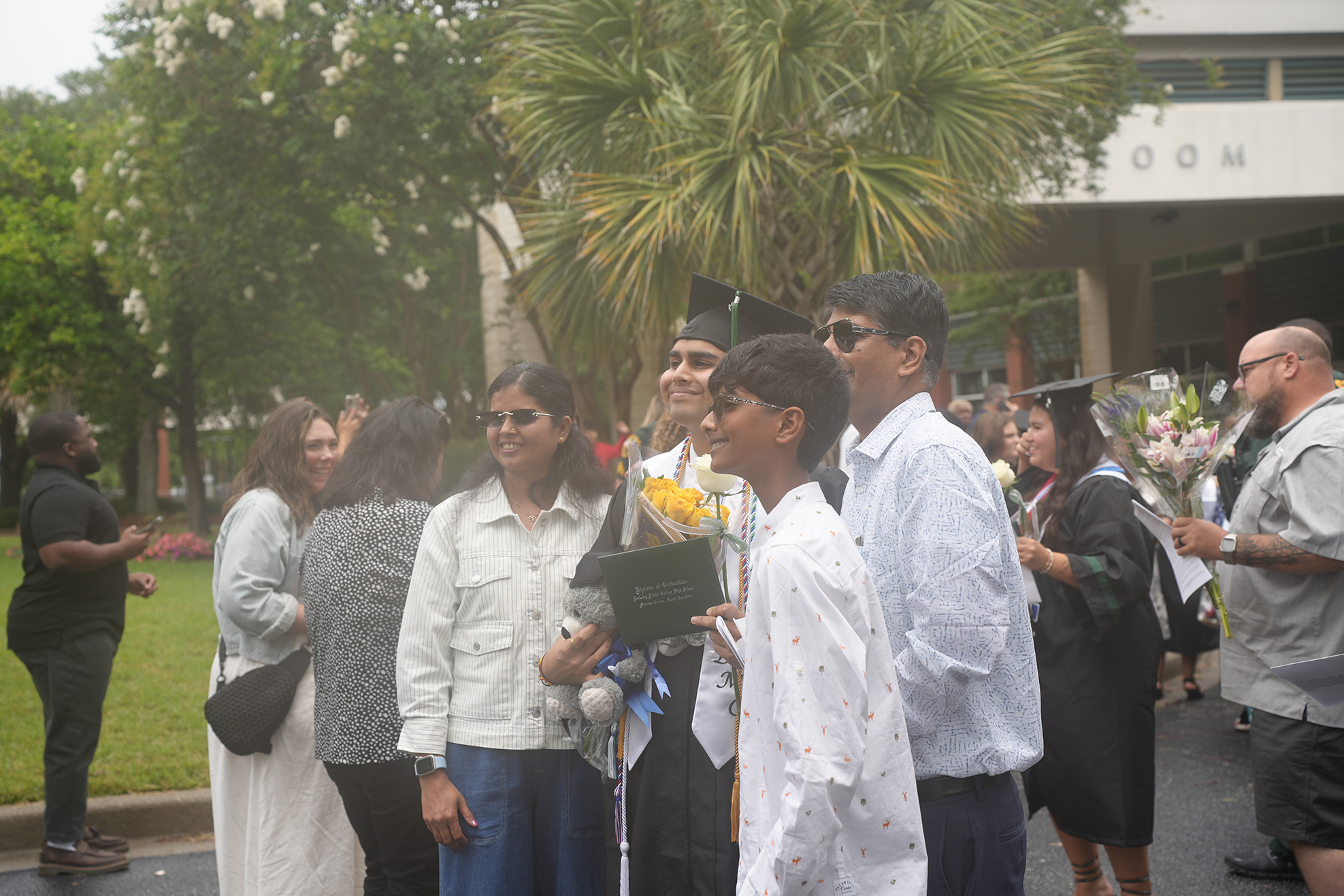 A group of people stand outdoors; some wear graduation gowns and caps. A woman holds a bouquet of flowers.