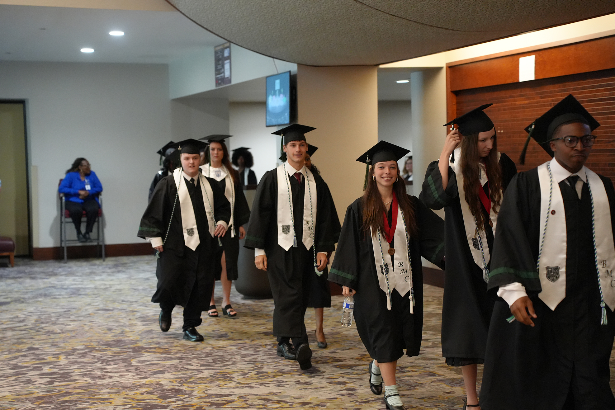 A group of people in academic gowns, some holding caps, walk along a carpeted corridor with arched ceilings.