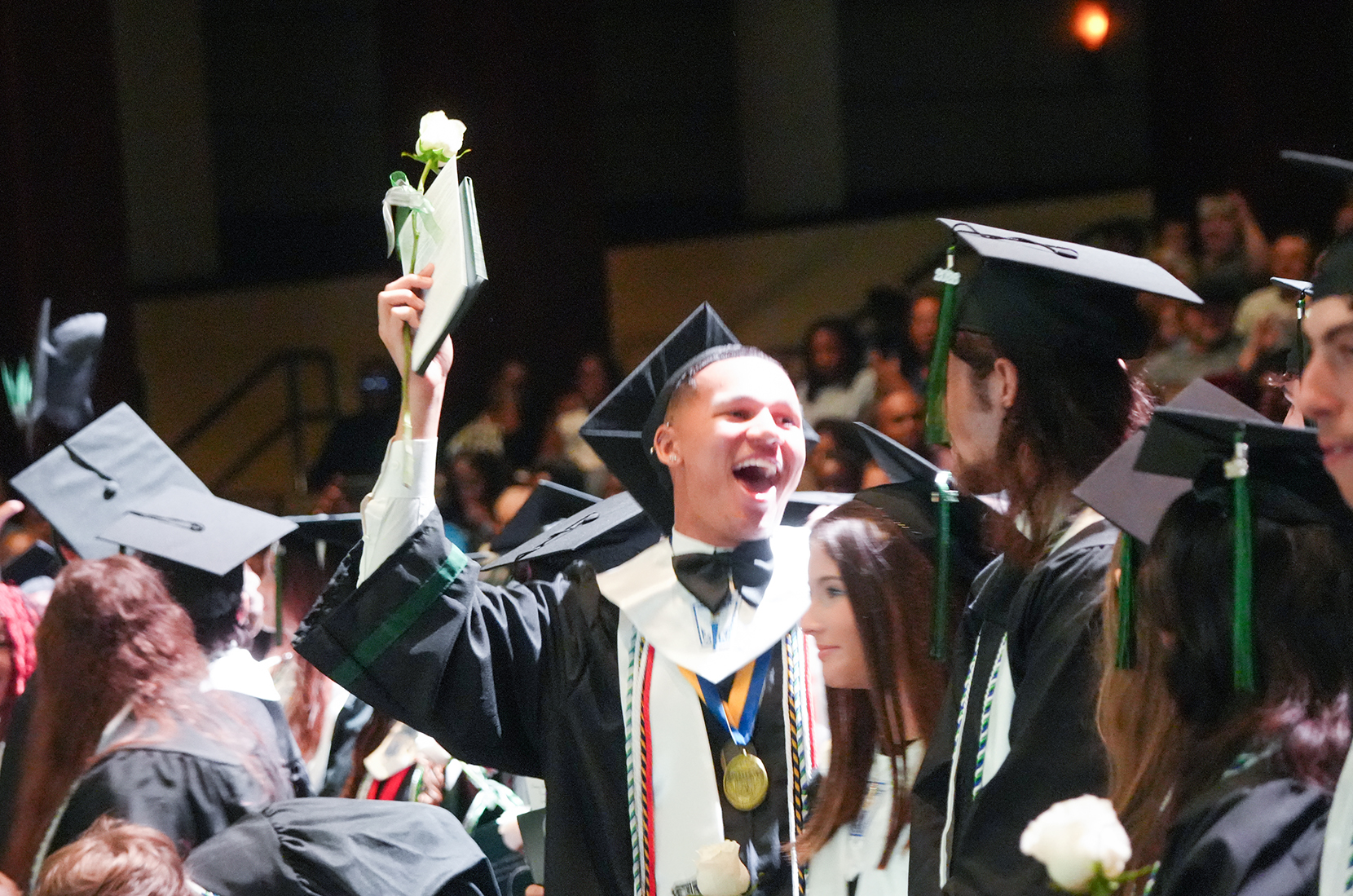 Graduates in black caps and gowns celebrating, one with a medal and flower, in a crowded auditorium.