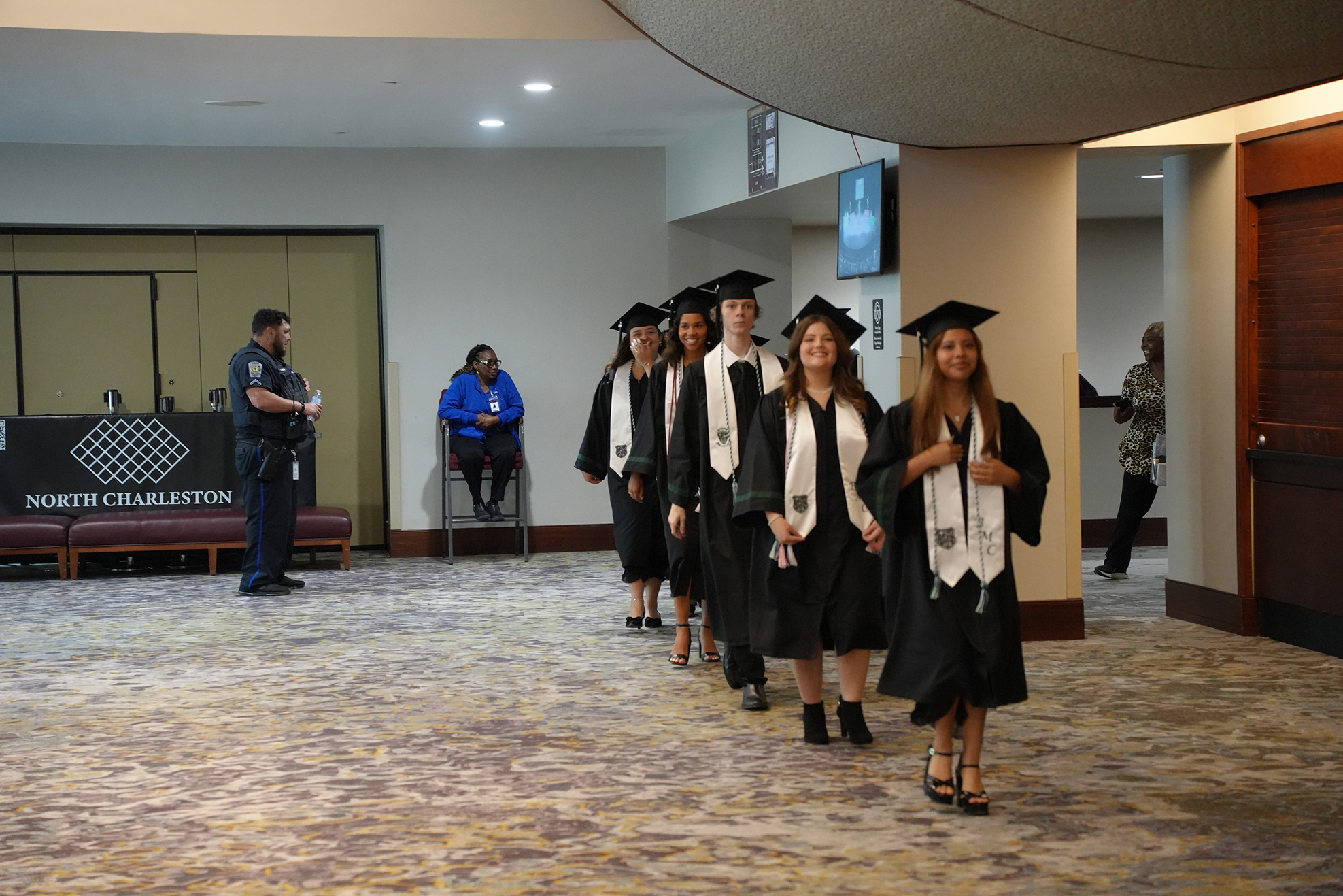 A group of graduates in caps and gowns walks down a hallway. A security officer stands nearby.