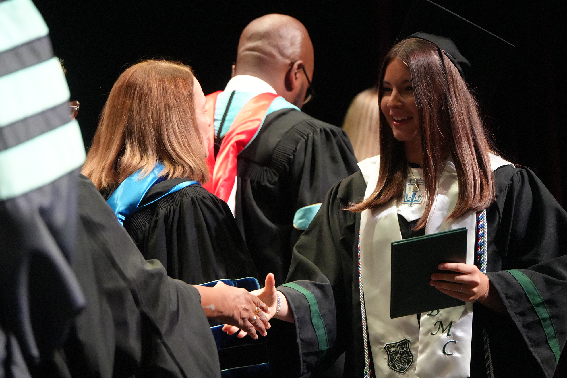 Graduates in black gowns and caps shake hands with each other in a row at an academic ceremony.