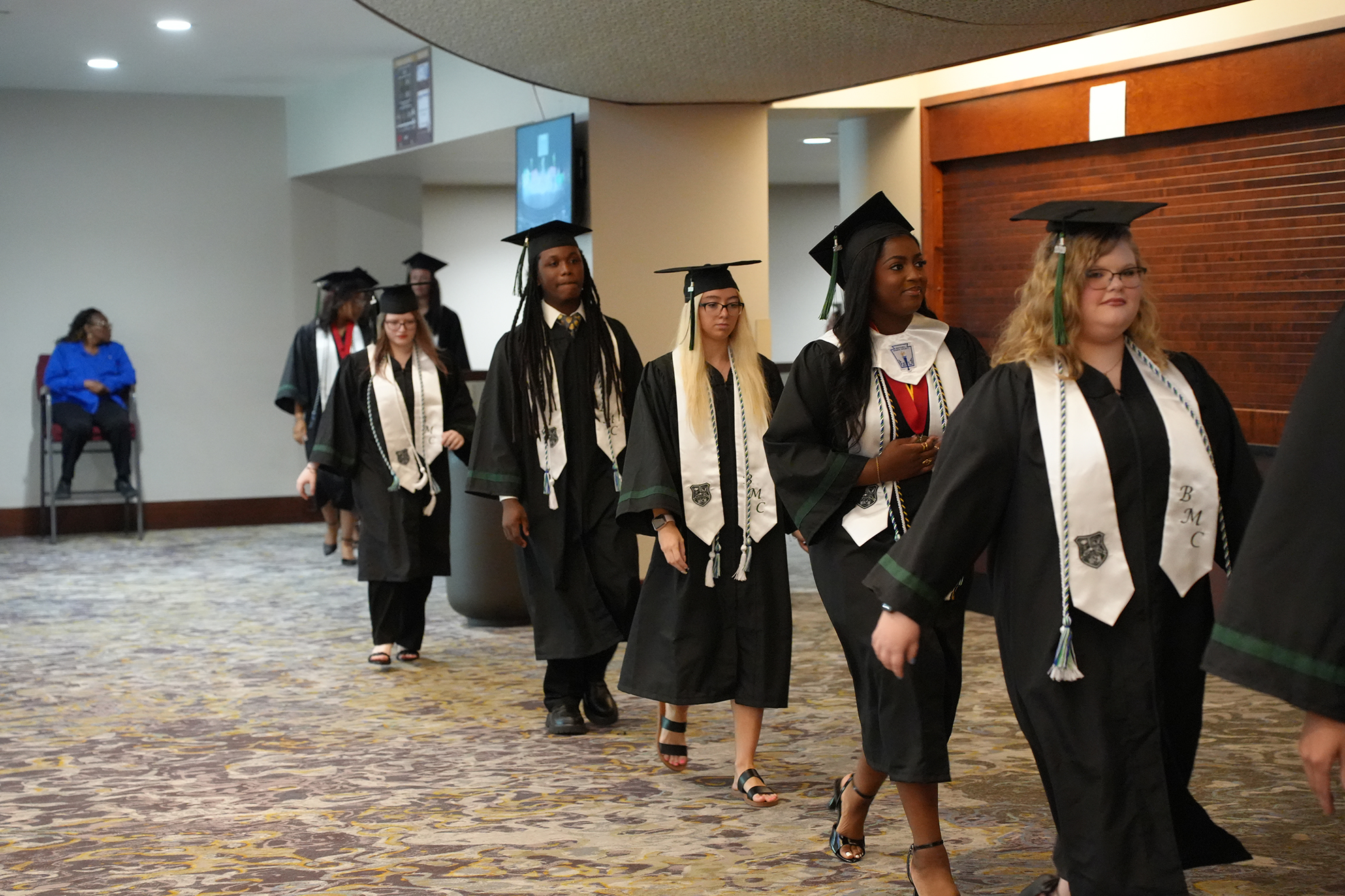 A group of six individuals in black gowns and hats walk through a hallway. A woman sits in a chair in the background.