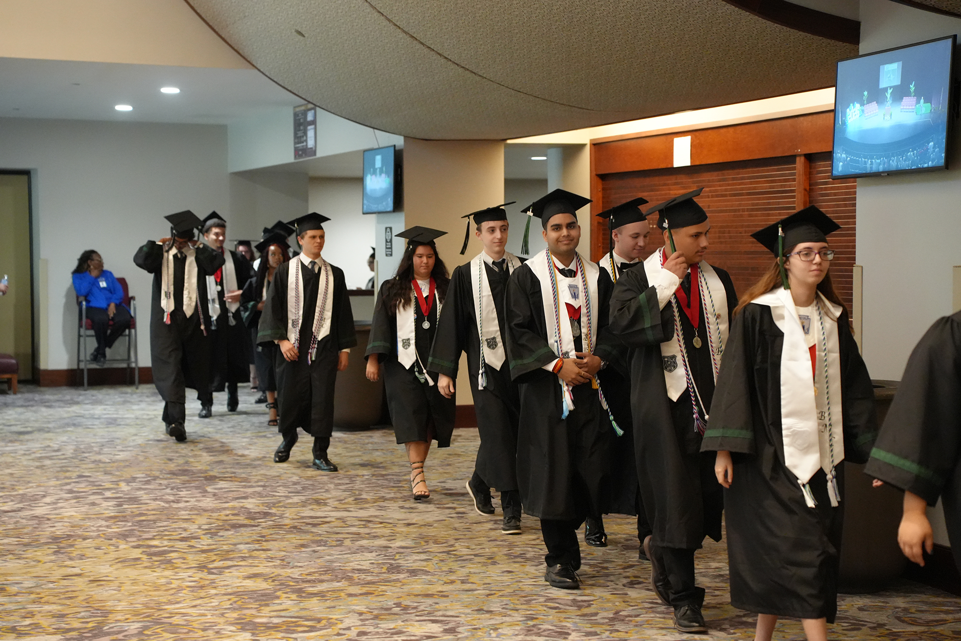 Graduates in black gowns and caps walk down a hallway; two women wear glasses, one has a white sash.