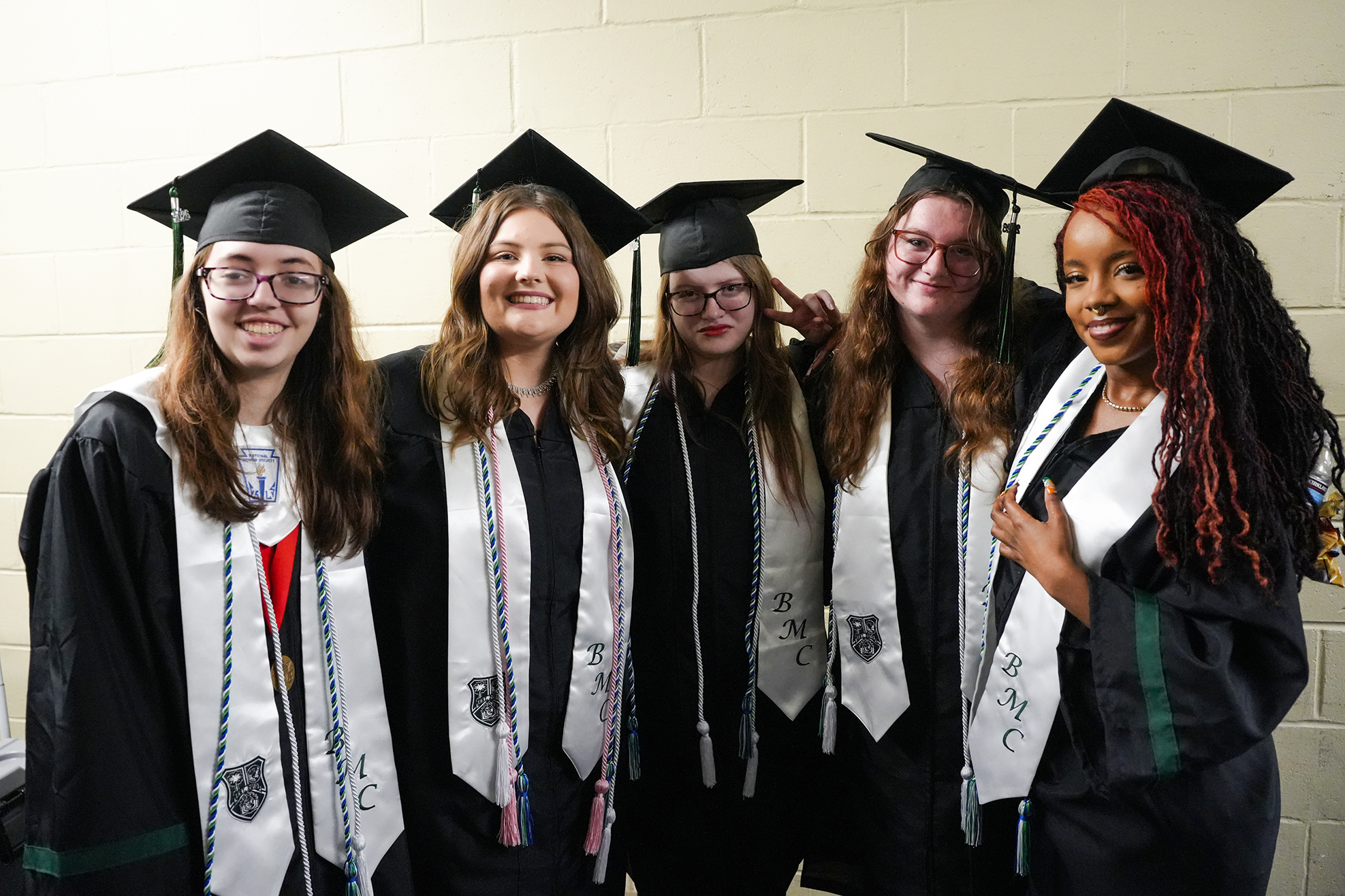 Five women in graduation gowns and caps standing against a plain white wall, smiling and posing for a photo.