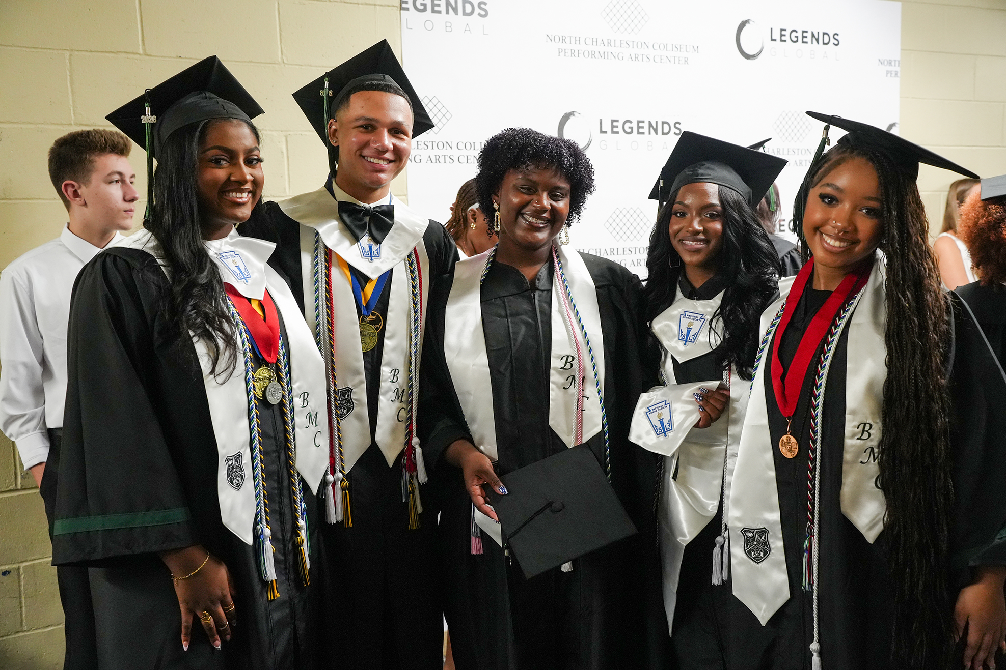Five people in black graduation gowns and caps stand together, smiling for a photo. Two are holding black folders, and the rest wear medals.