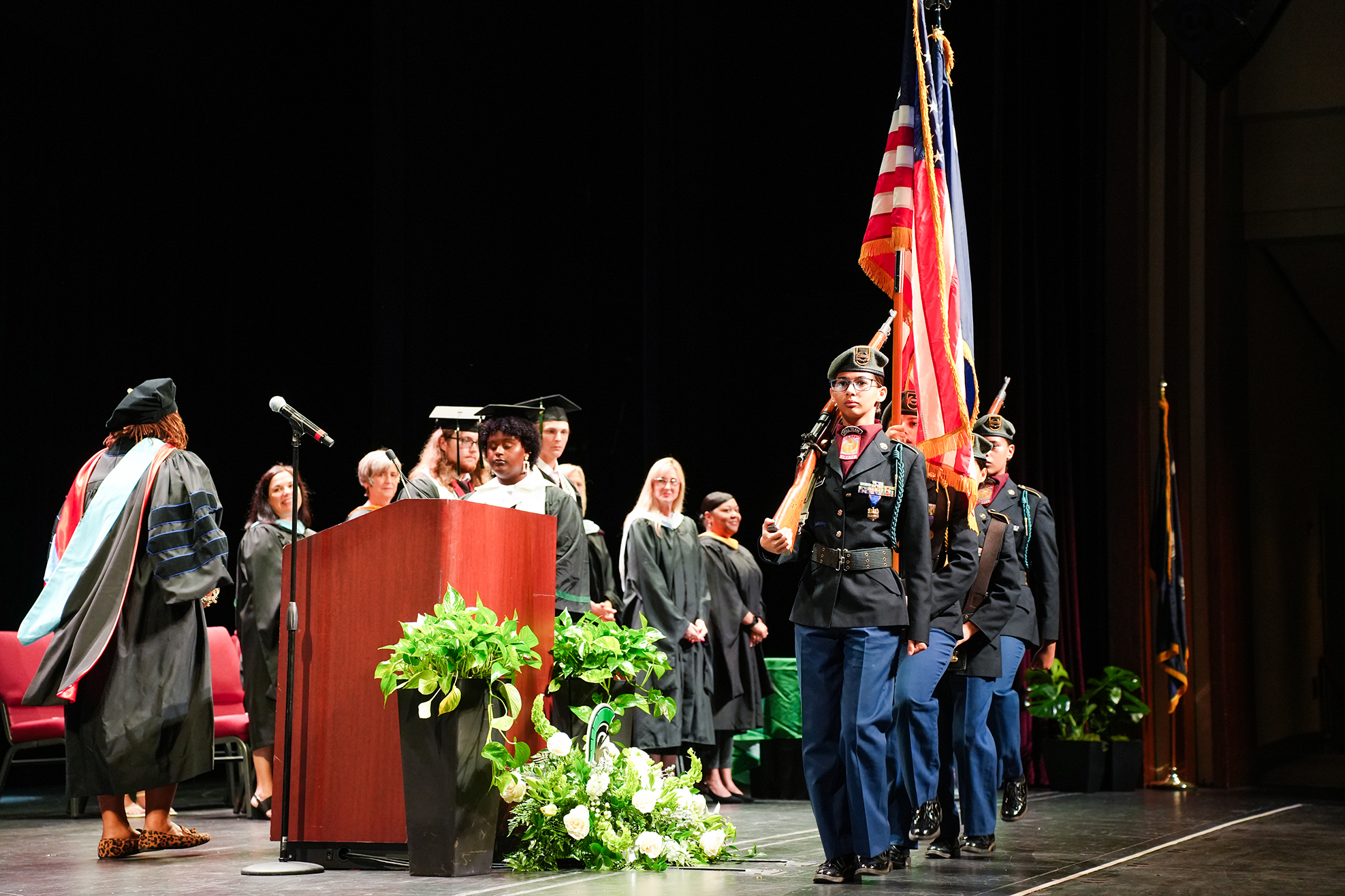 A podium on stage has graduates in caps and gowns. A soldier carries a flag. Some people stand near the podium.