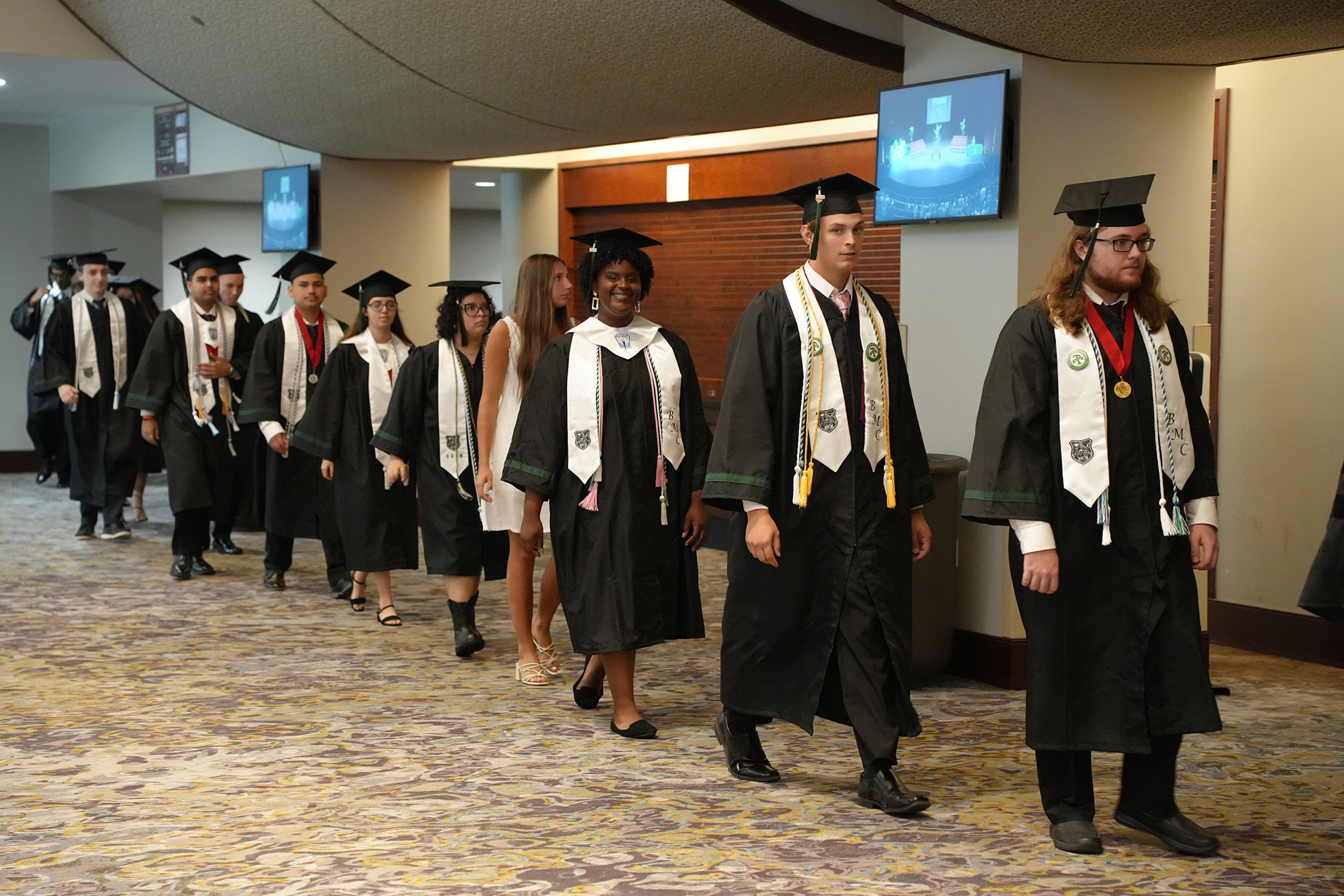 Graduates in black gowns and caps walk in a line down a carpeted hallway, some wearing medals.