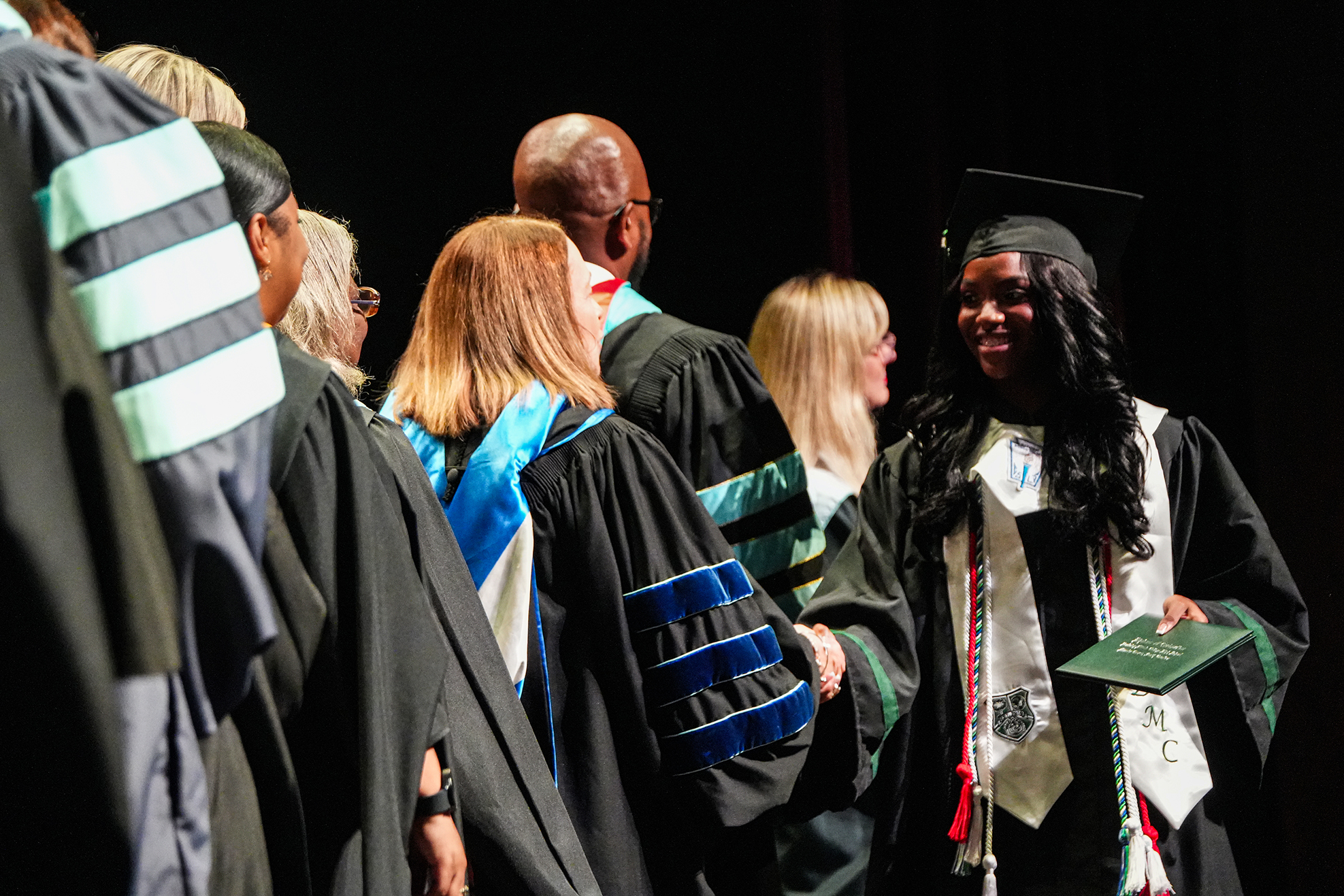 A crowd of people in academic regalia at an event, a woman in a graduation gown holds a diploma.