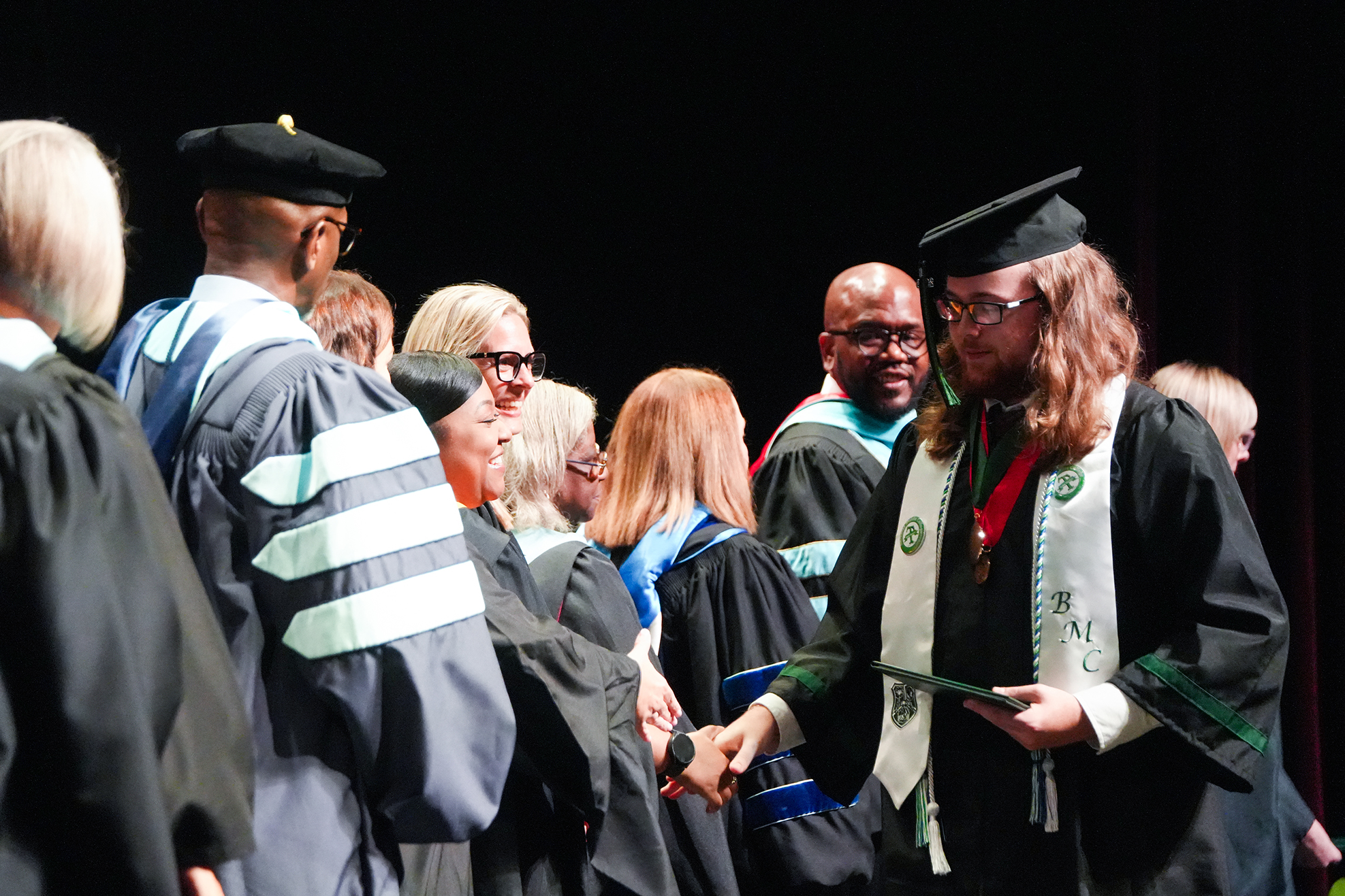 A group of people in academic regalia stand at an event. One person shakes hands with another while holding an award.