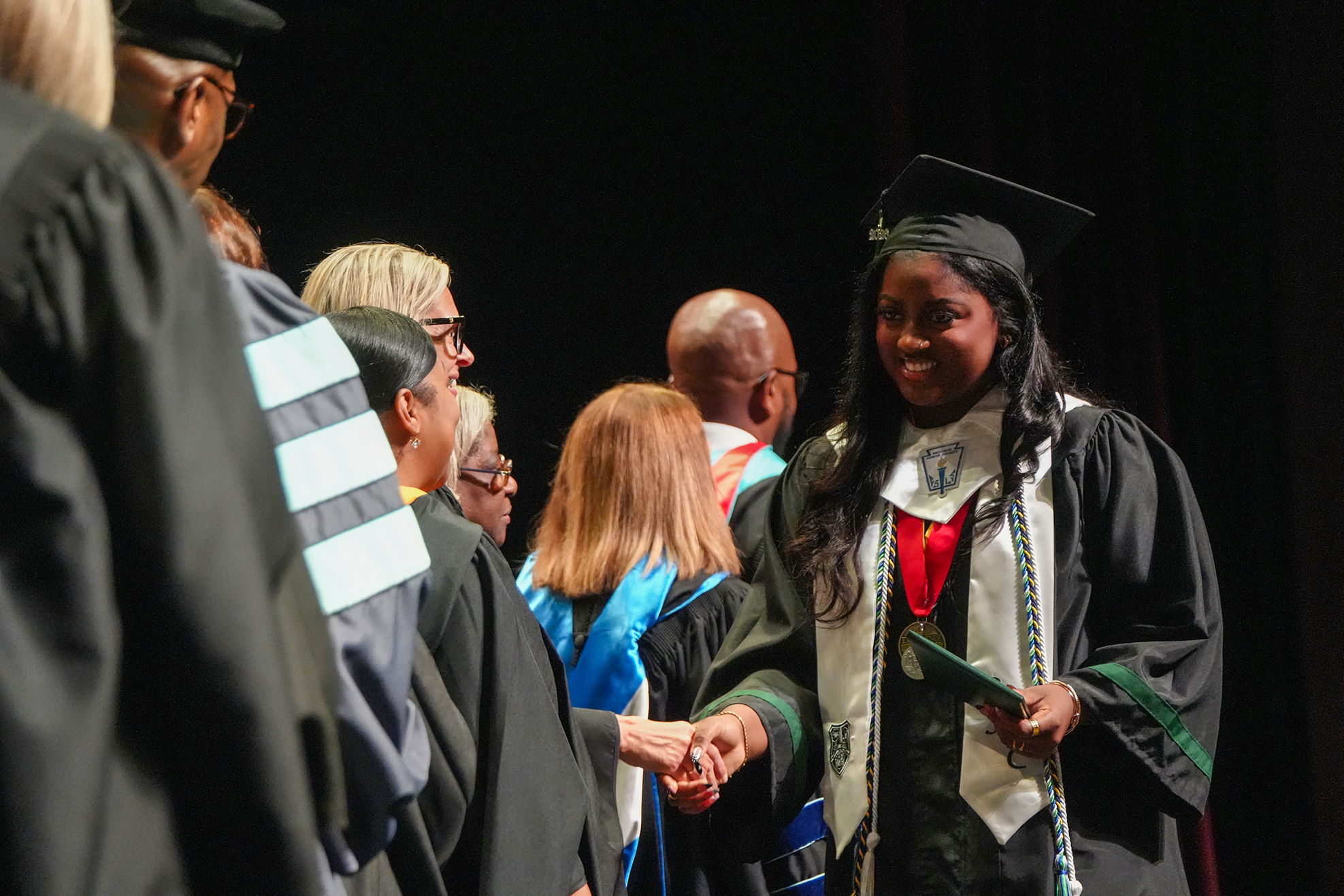A woman in a graduation gown shakes hands with others, who are also wearing graduation gowns and caps.