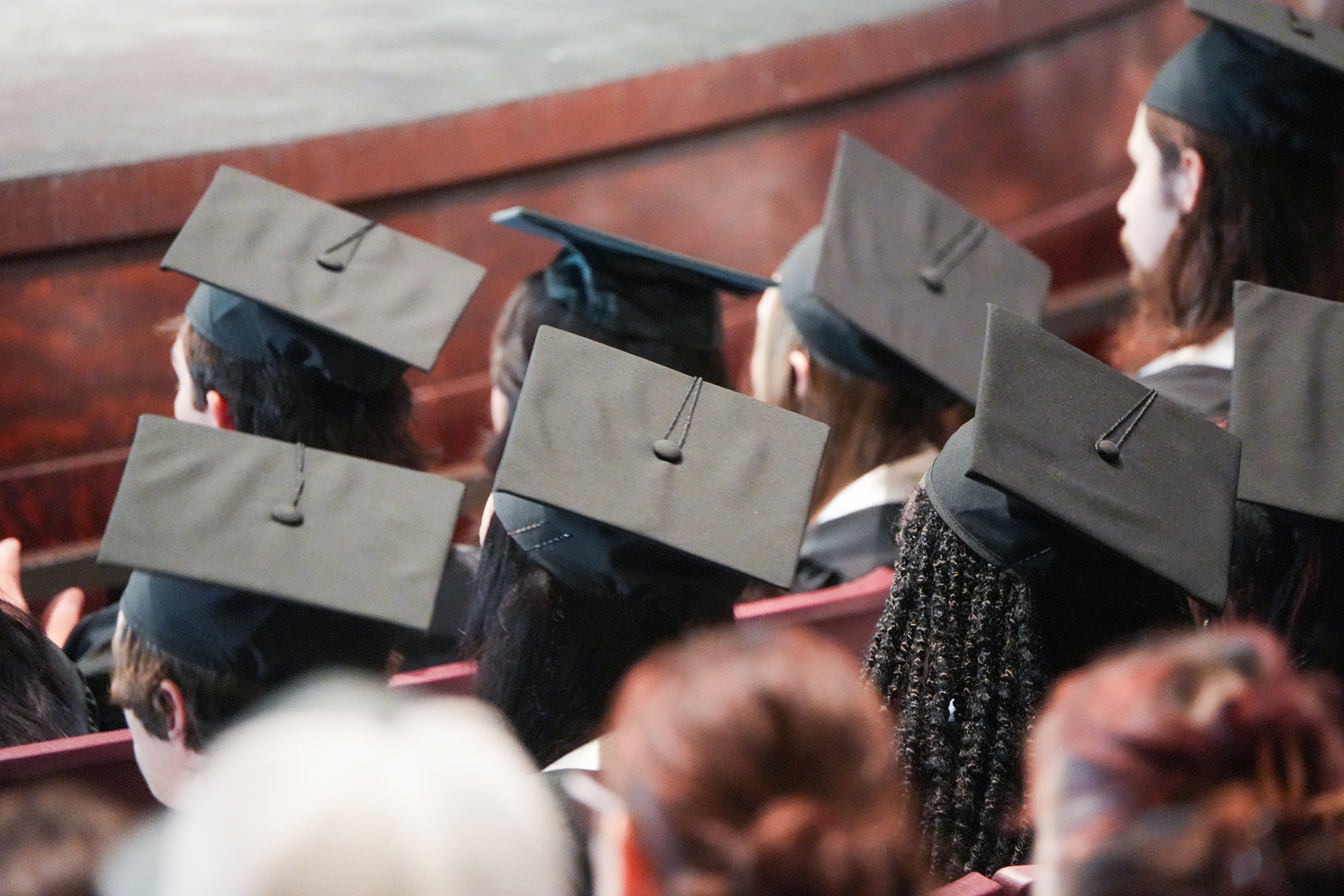 A group of individuals wearing graduation caps, attending a ceremony in a venue with wooden and red seating.