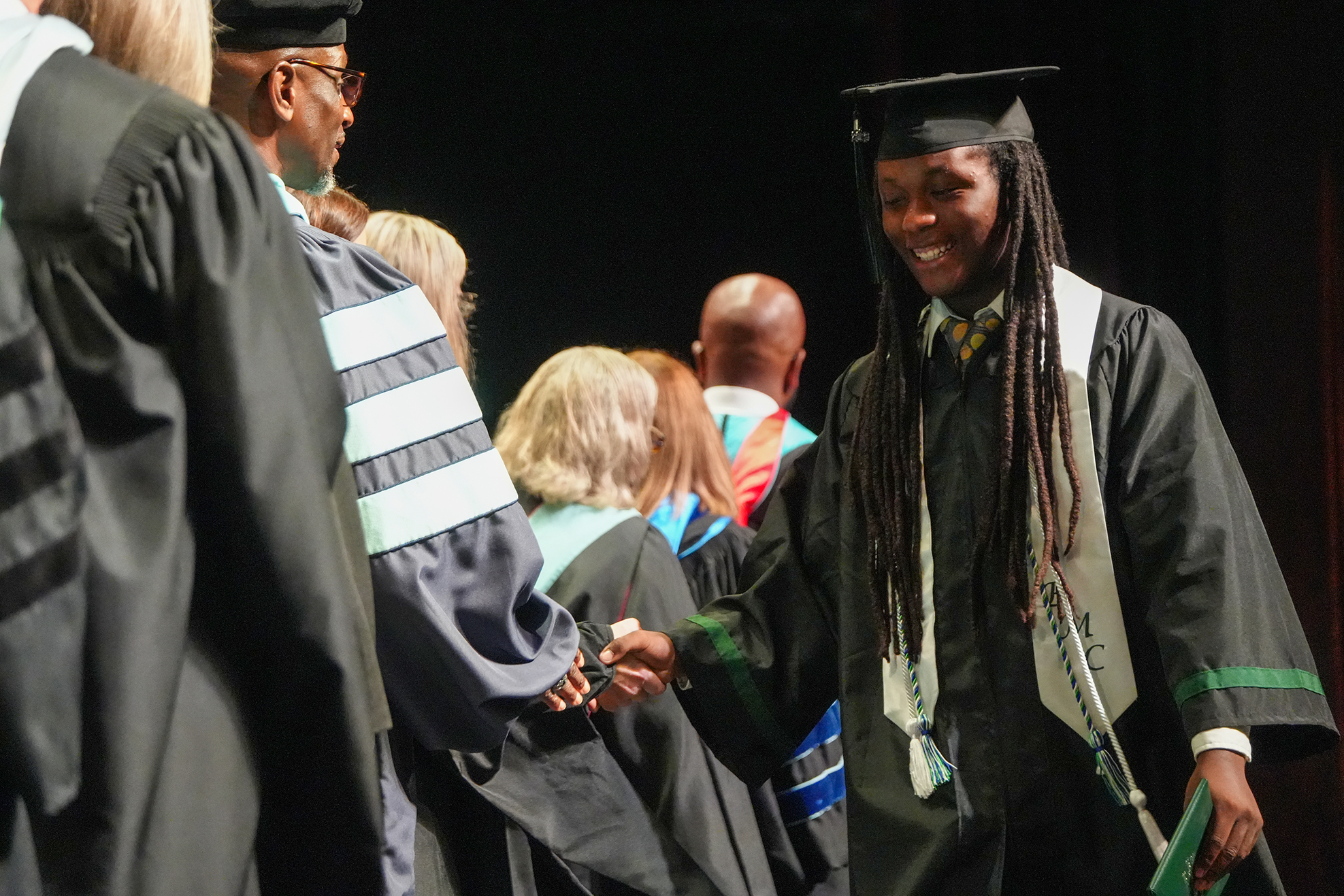 A person in a graduation gown shakes hands with another person at a ceremony. Behind them, others in graduation gowns are present.