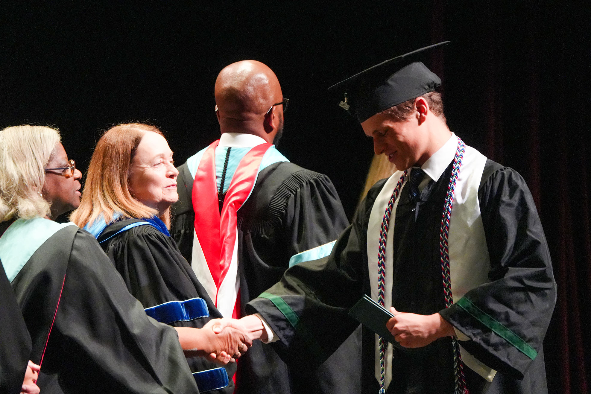 A group of individuals in academic regalia stand together. A man in a graduation cap shakes hands with a woman.
