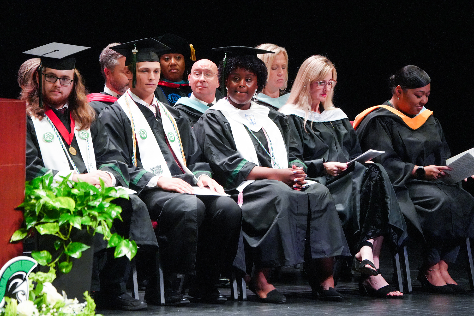 Graduates in academic regalia seated on stage, facing forward, during a ceremony. Some hold papers.