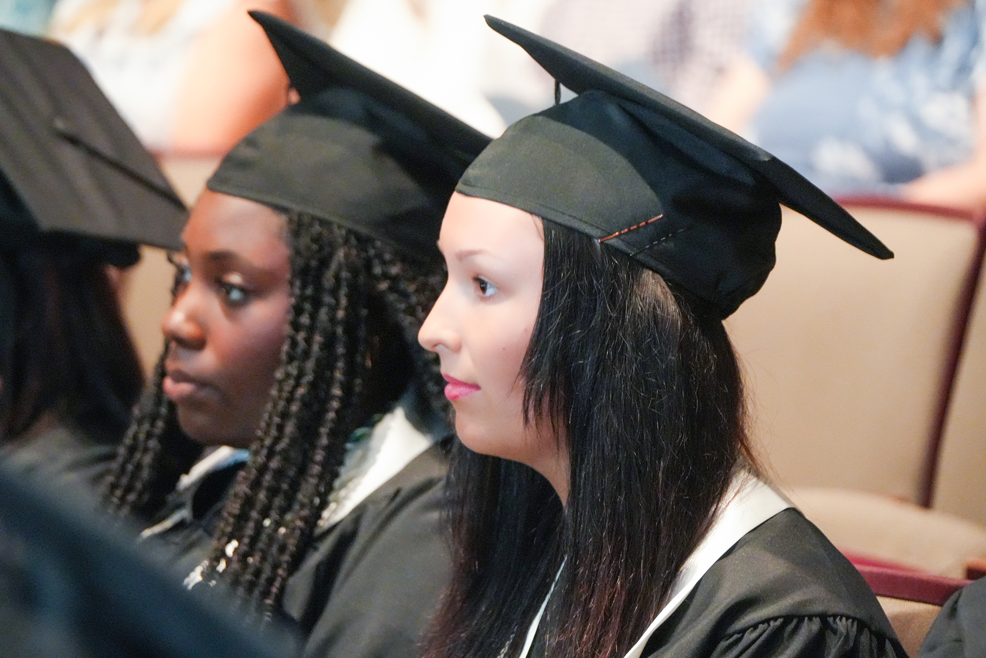 Two women in graduation attire with black caps and gowns, one with braids, attend a ceremony.
