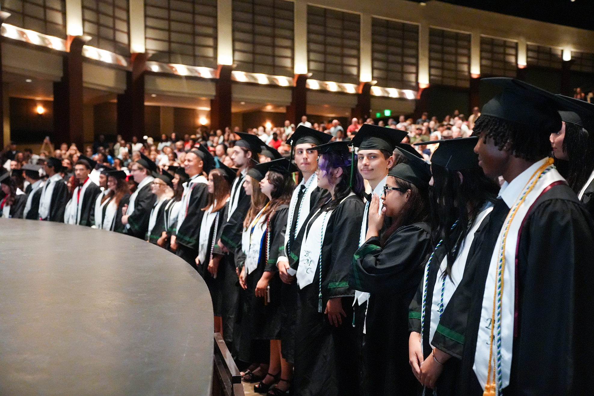 A group of students in graduation robes and caps stand in a line on stage in a large hall.