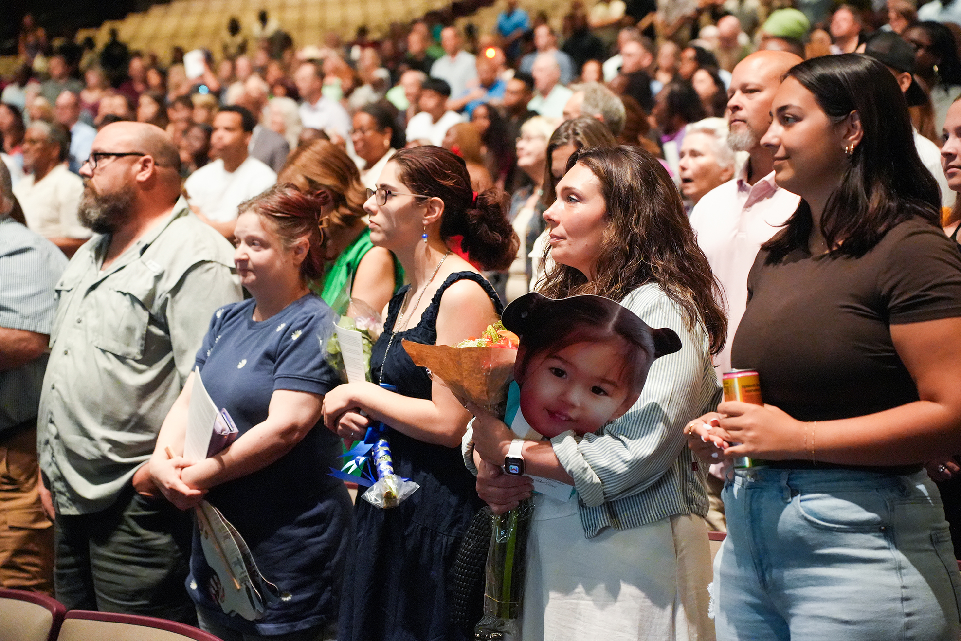 A crowd in a large room; some people hold flowers and some hold cutouts of a baby.