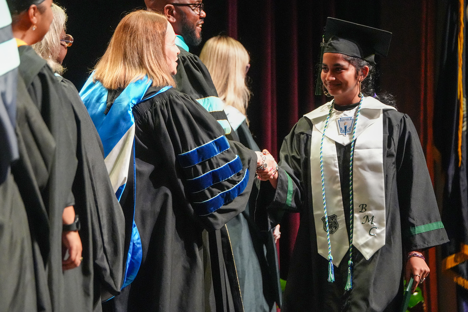 Graduates in academic regalia stand on stage; one shakes hands with another. A woman wears a cap and gown.
