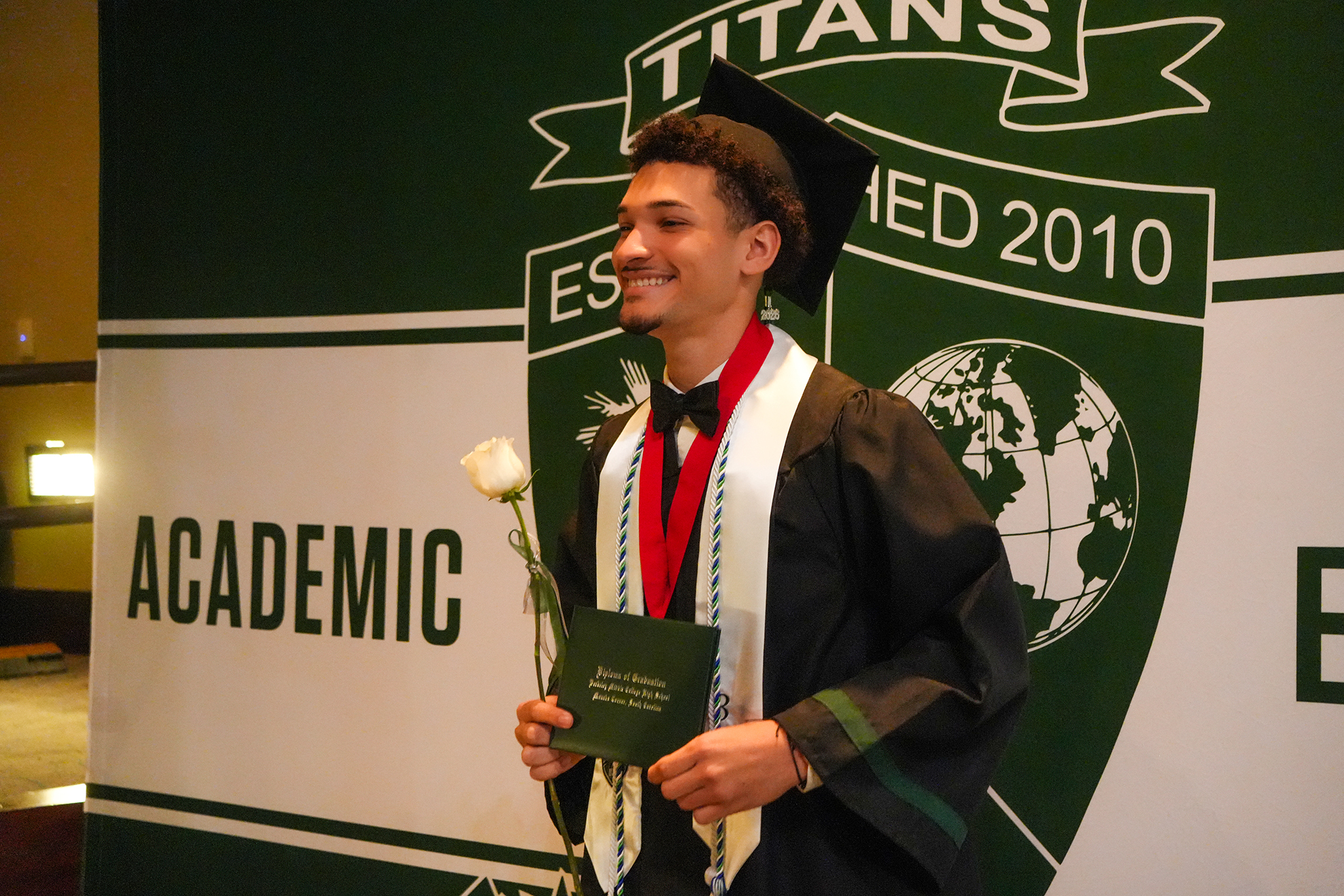 A male student stands in a cap and gown holding a white rose, smiling, in front of a decorative backdrop.