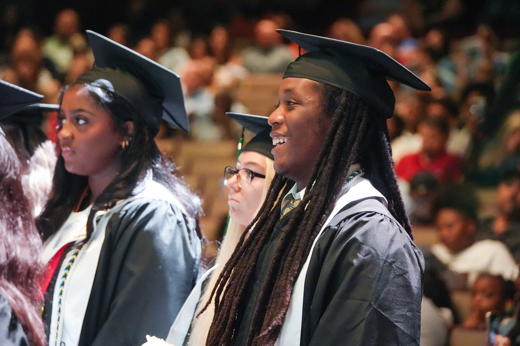 Two individuals in black graduation gowns stand in a row, facing a crowd in a large room.