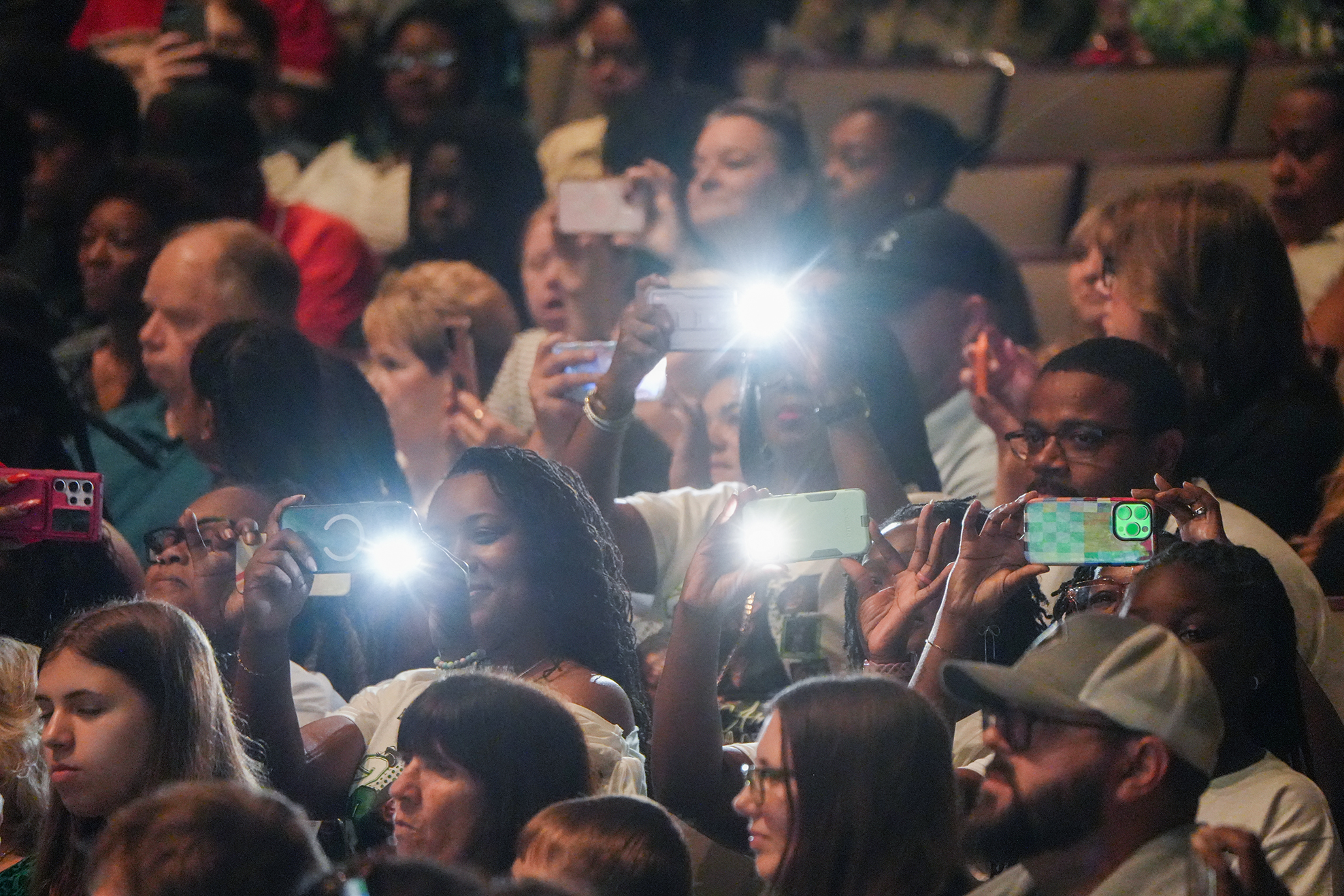 A crowd of people in a theater, many holding up flashlights and phones, capturing a moment.