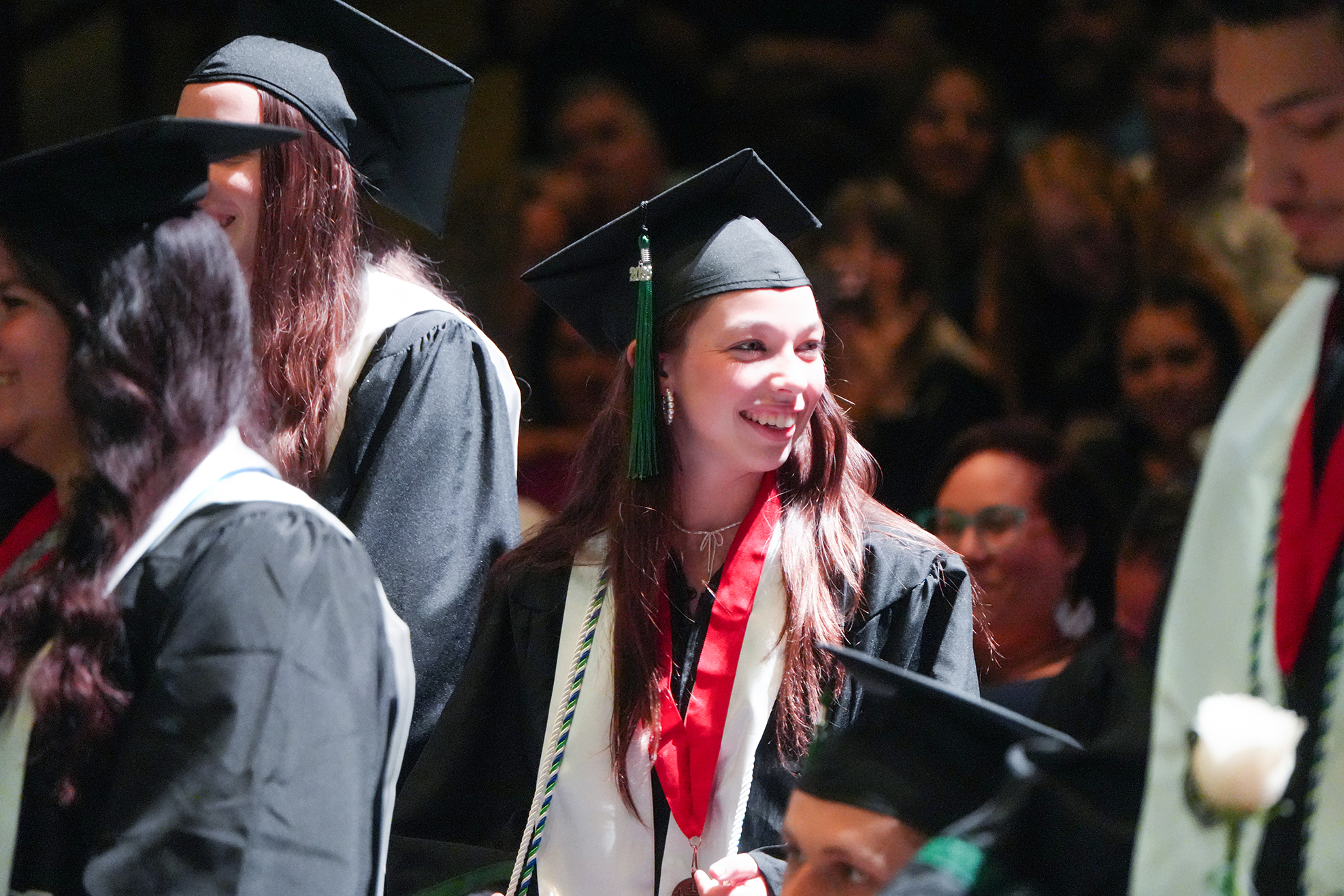 Graduates in black gowns and caps, one with a red cord, smiling at a ceremony.