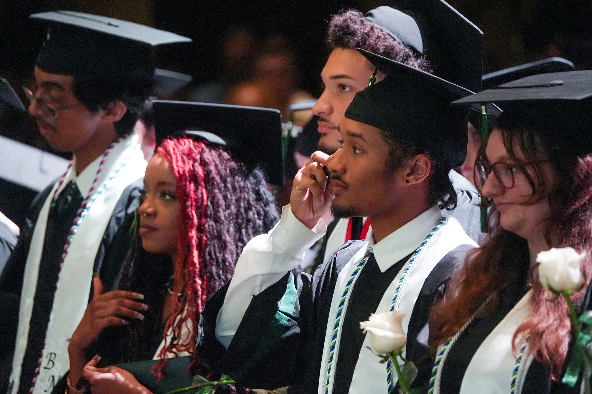 People in caps and gowns at a graduation ceremony. One person holds a rose.