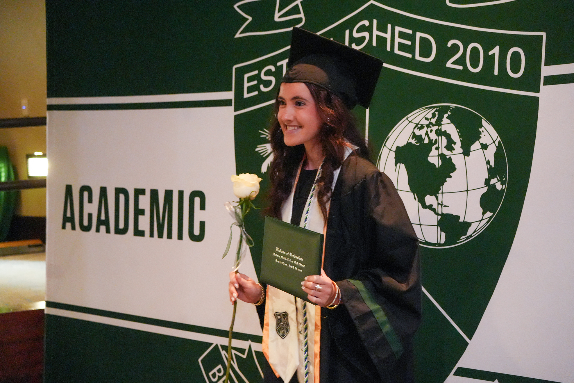 Person in graduation attire holds a rose and diploma in front of a green banner.