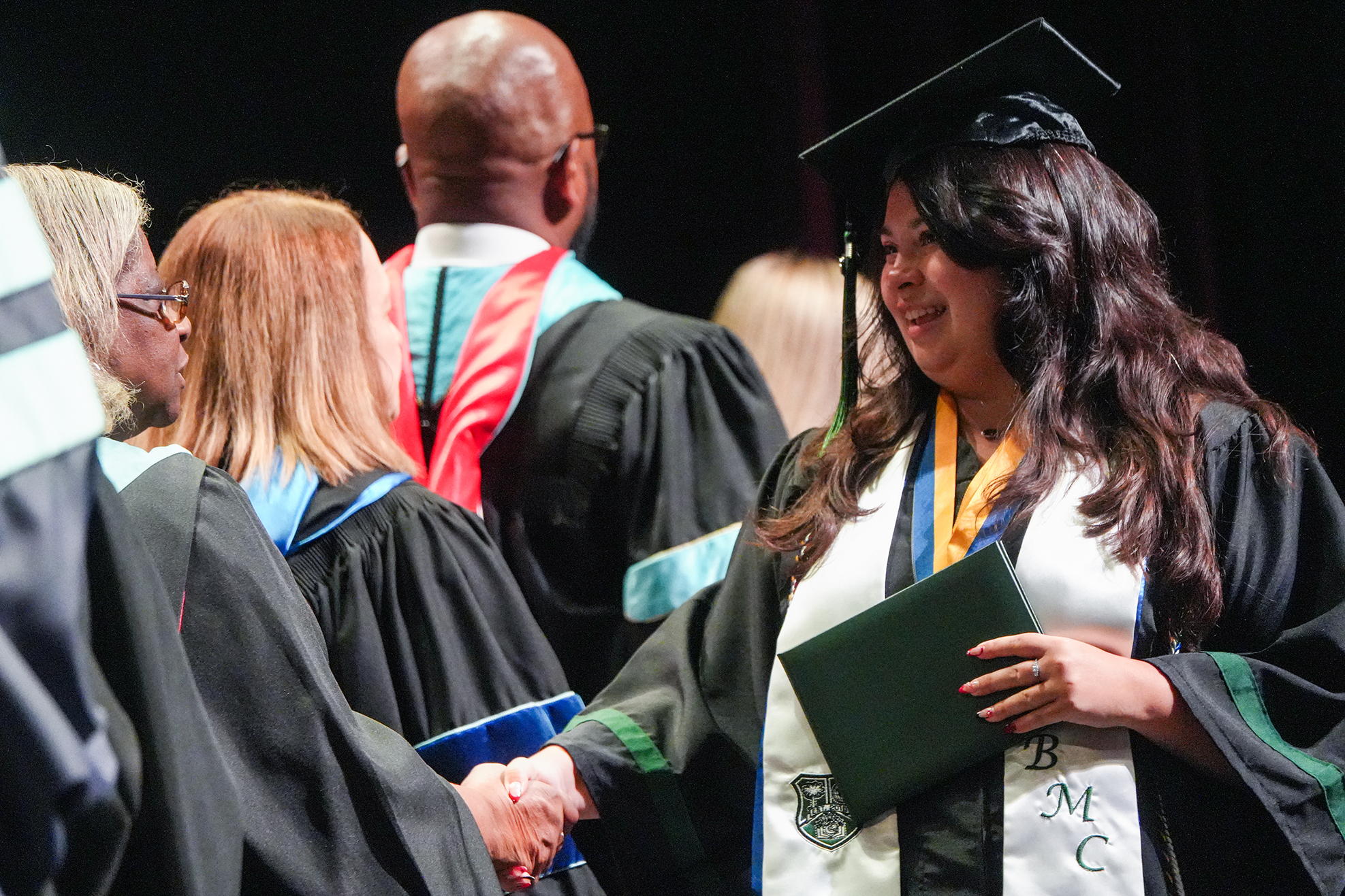 Woman in a graduation gown and cap, holding a diploma, stands before others in academic attire.