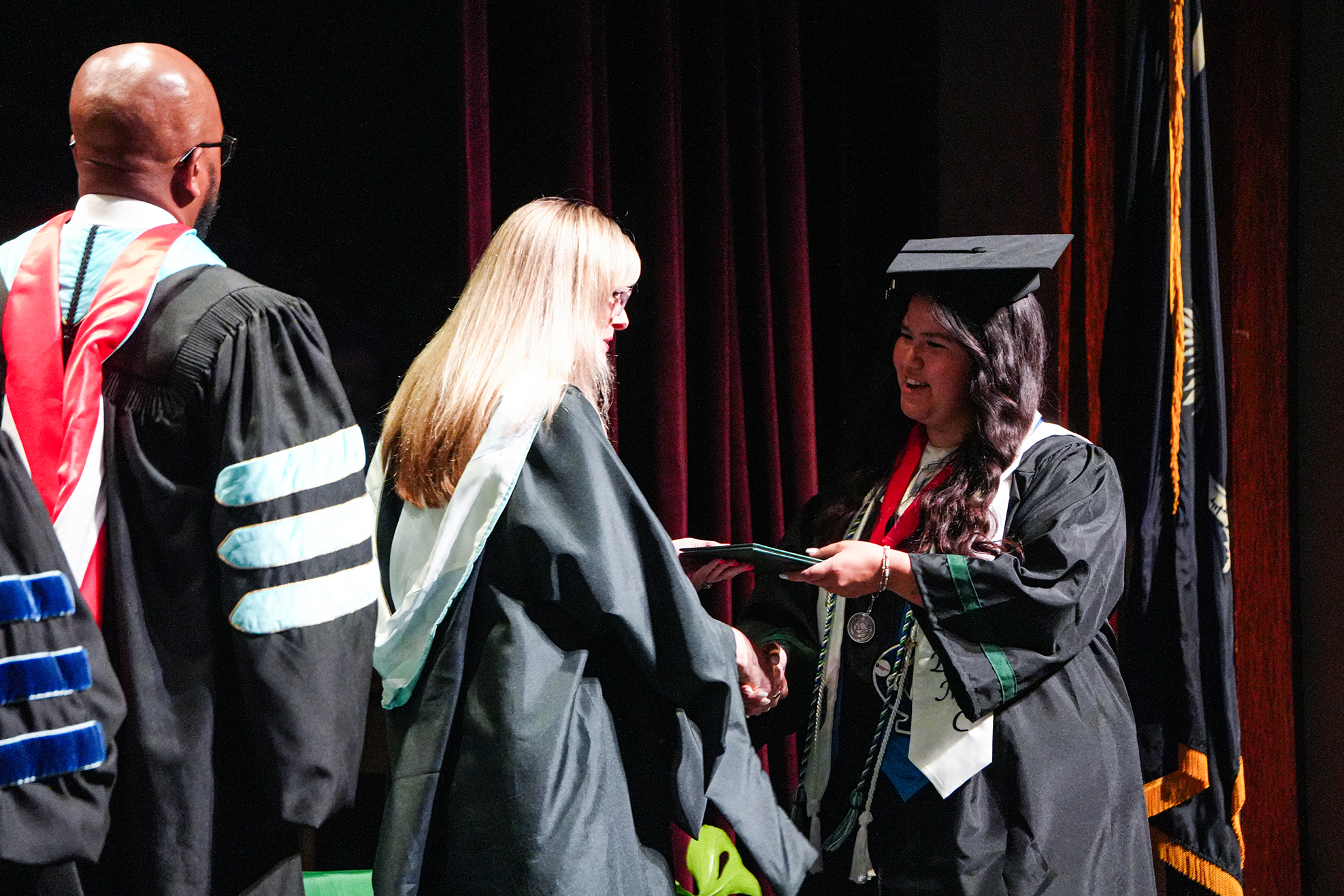 A woman in a graduation gown shakes hands with another woman. They are standing on a stage.