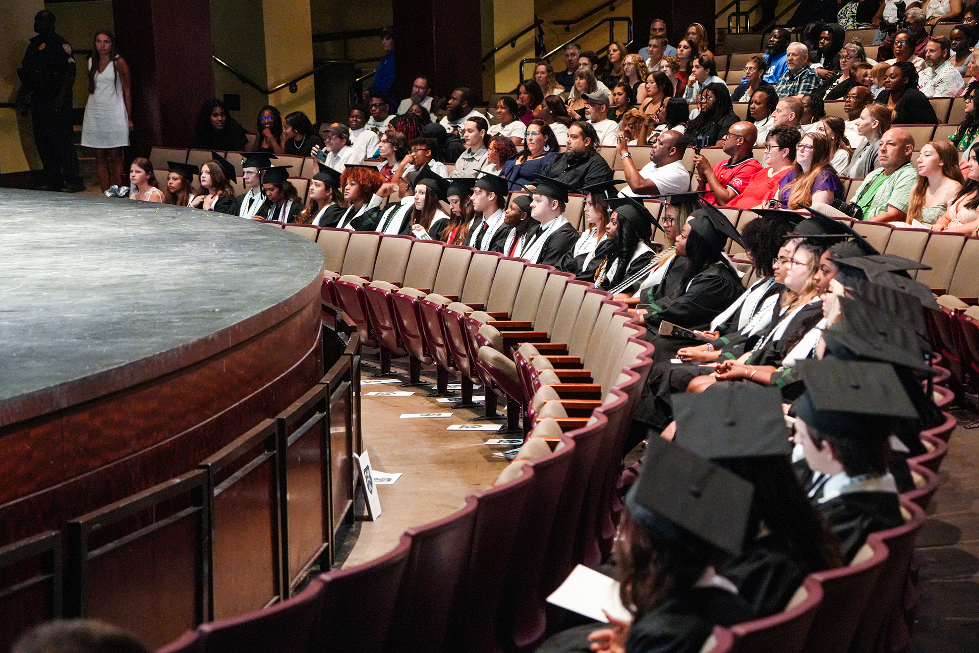 Many graduates in caps and gowns sit in rows facing a stage, where a woman in a white dress stands.