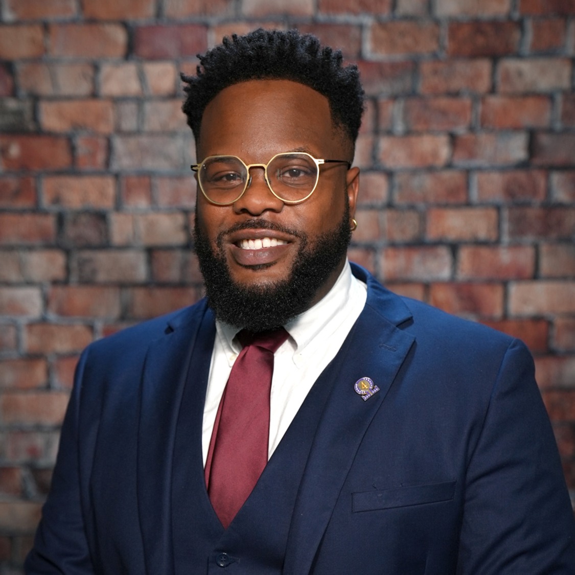 A man in a blue suit, red tie, and gold-framed glasses stands smiling. Behind him is a red brick wall.