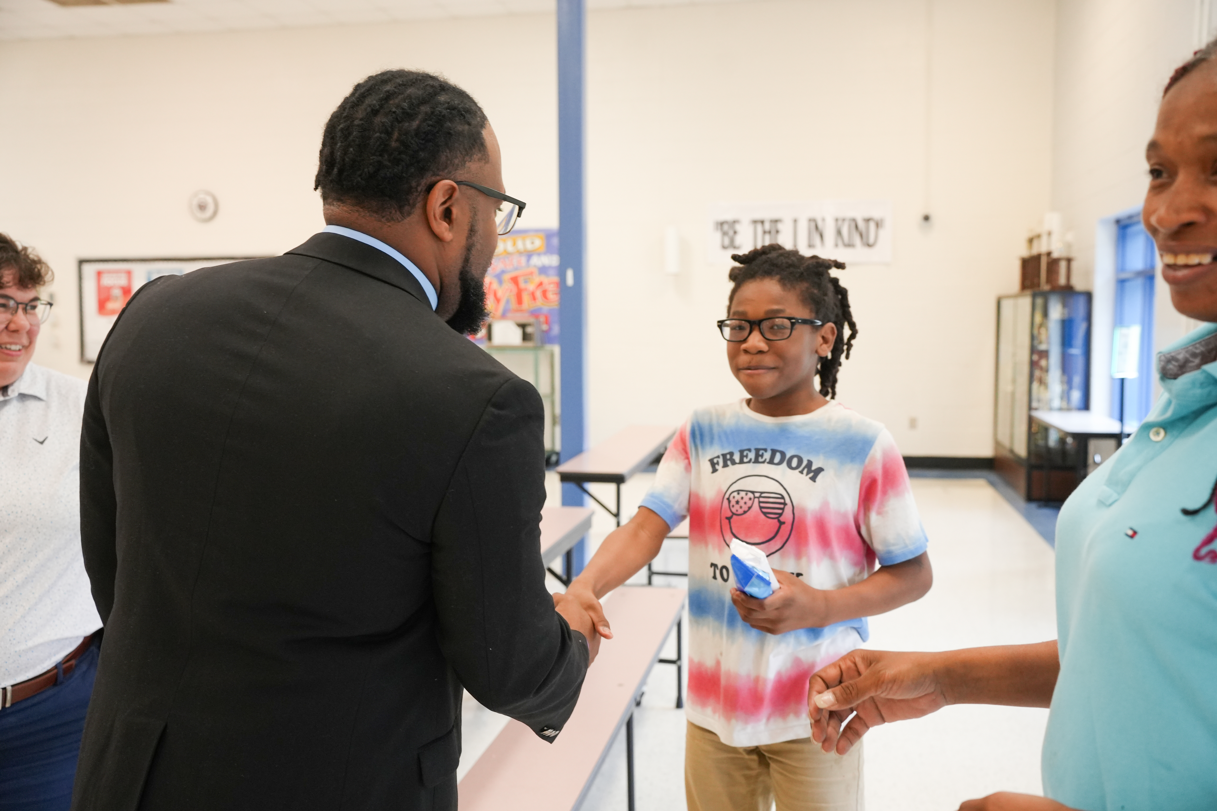 Four people in a classroom. A man in a suit shakes hands with a girl. Another adult holds a small object.