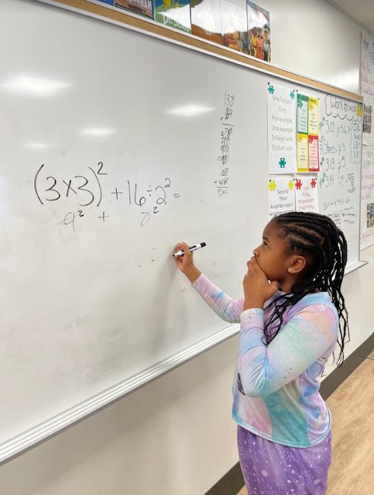 A girl works to solve a complicated multiplication multi-step problem on the board.