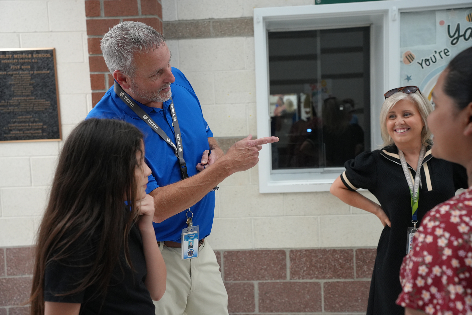 A man in a blue shirt points at a girl in a black shirt; a woman in a black dress smiles nearby.