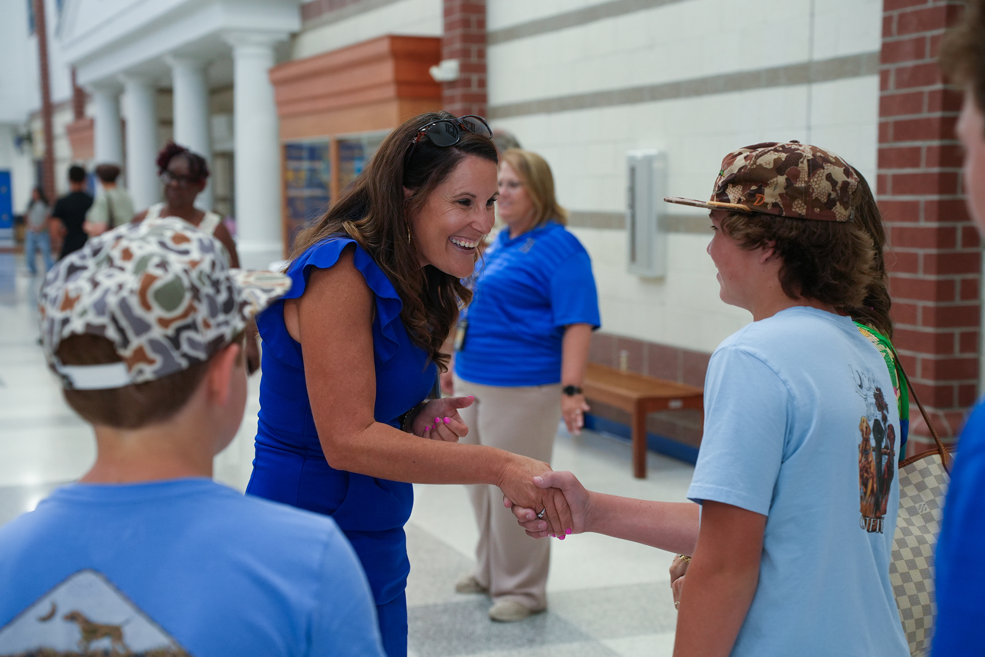 A woman in a blue outfit smiles while shaking hands with two students. The students wear blue t-shirts and caps.