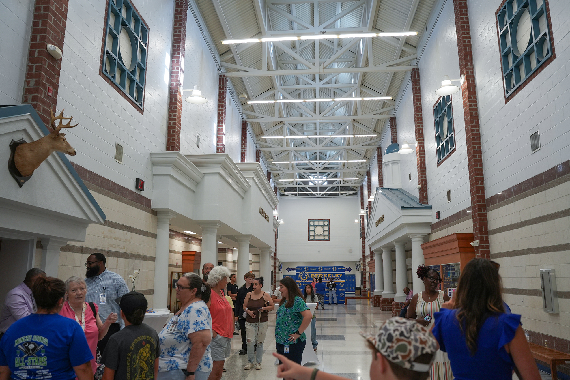 A hallway in a building with white walls, metal beams, and glass windows. People stand and walk in groups.