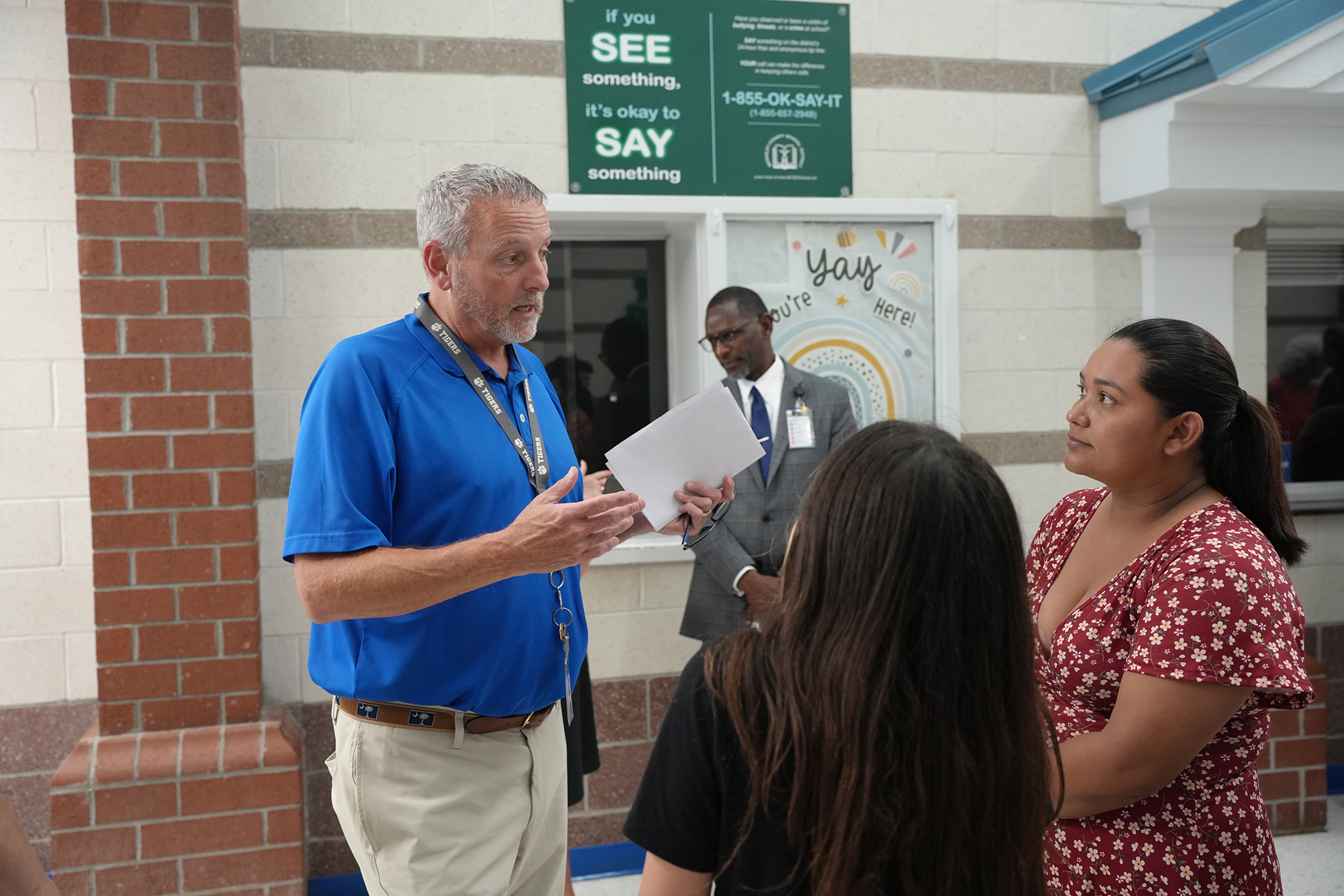 A man in a blue shirt and khakis talks with two women in front of a building.