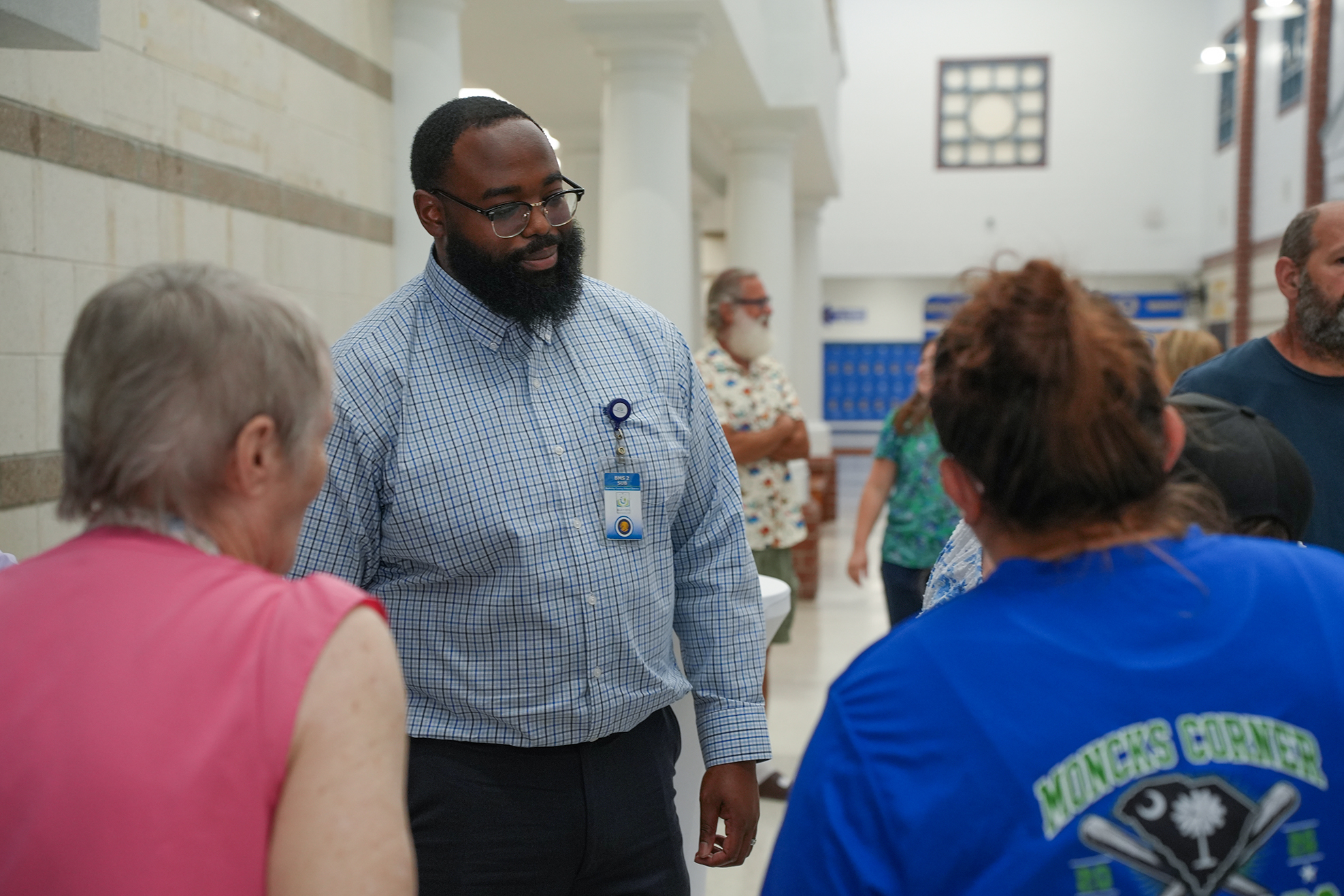 A man with a beard and glasses speaks with people. One person wears a blue shirt and a cap.